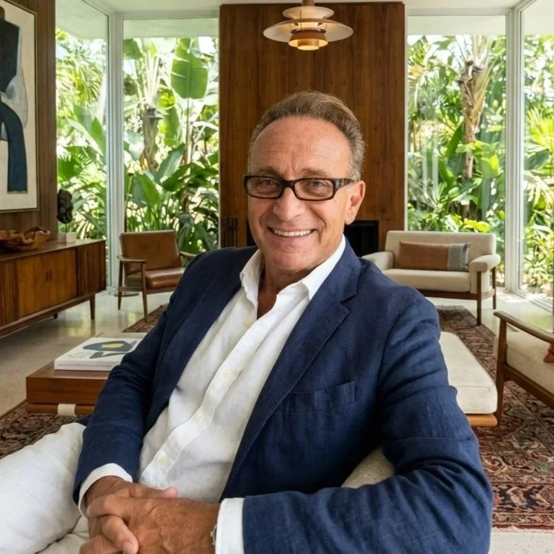 A man smiling and sitting in a modern, well-lit living room with large glass windows and tropical greenery outside. The room features wooden furniture, a patterned rug, and a unique ceiling light fixture.