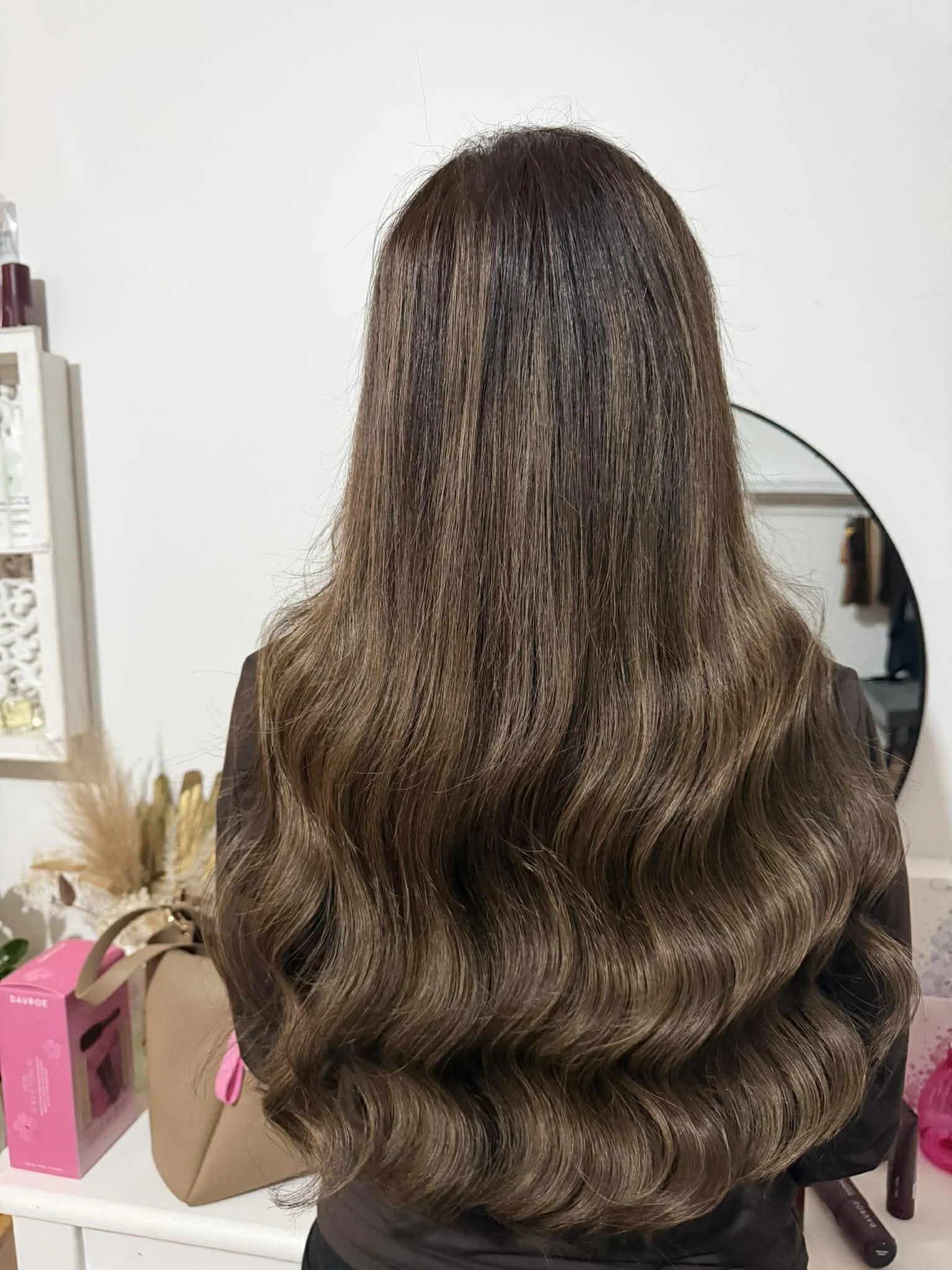Back view of a woman with long, wavy, brown hair with highlights, in a salon or dressing area, standing in front of a mirror.
