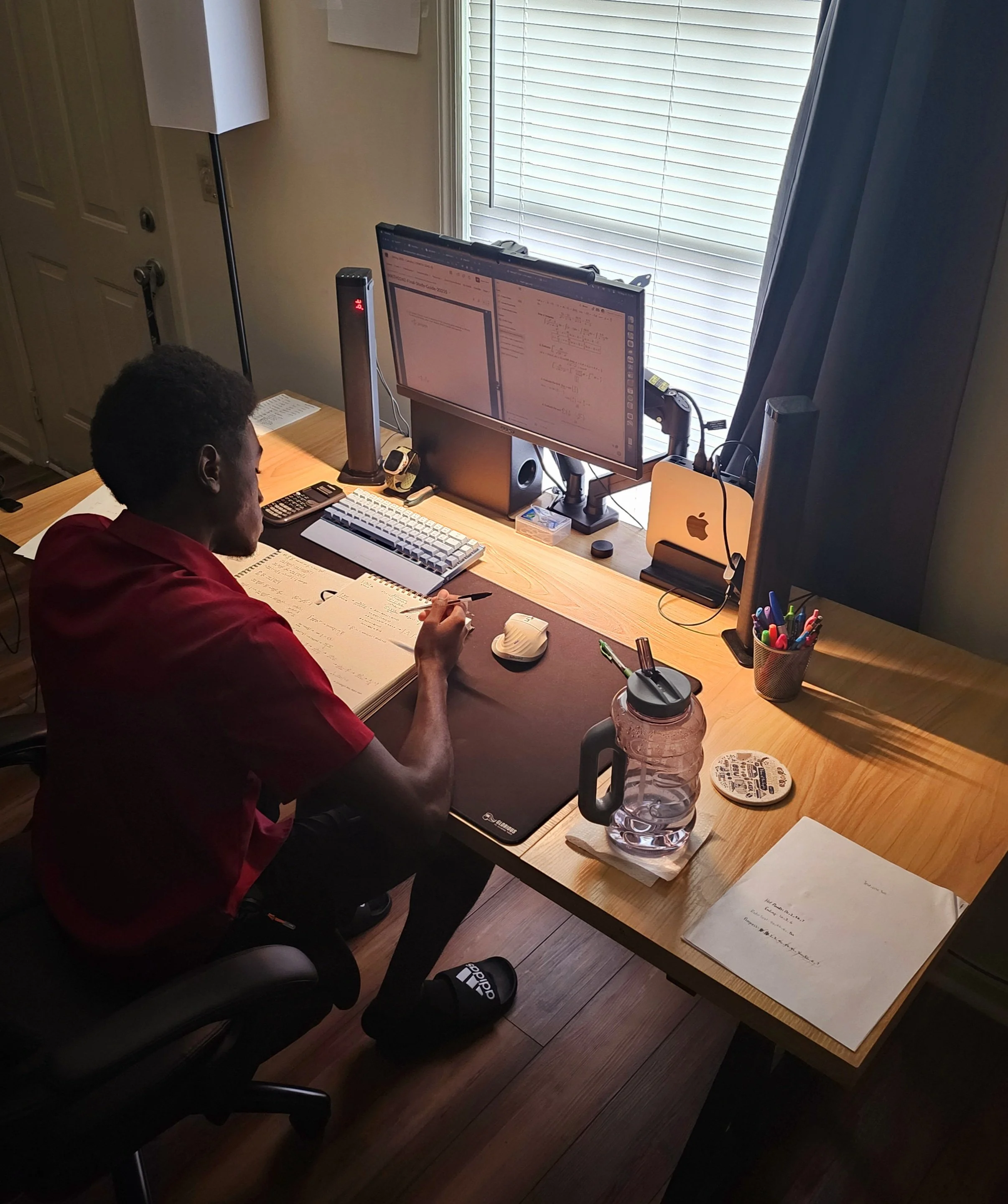 A person sitting at a desk working on a computer with dual monitors, notes, and cups inside a room with a window covered by blinds.