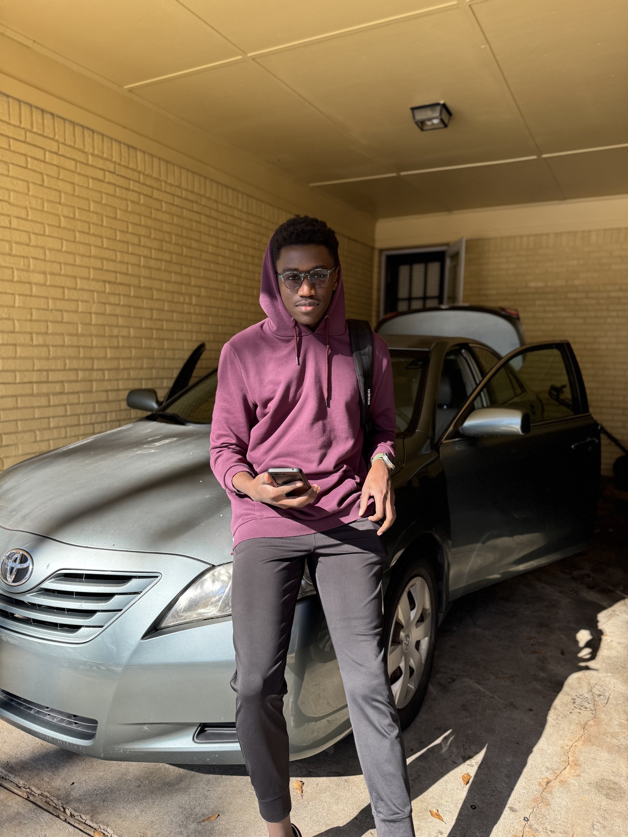 A young man wearing a maroon hoodie, sunglasses, and gray pants leaning against a silver car parked outside a building with yellow brick walls. He's holding a smartphone and has a black backpack on his shoulder.