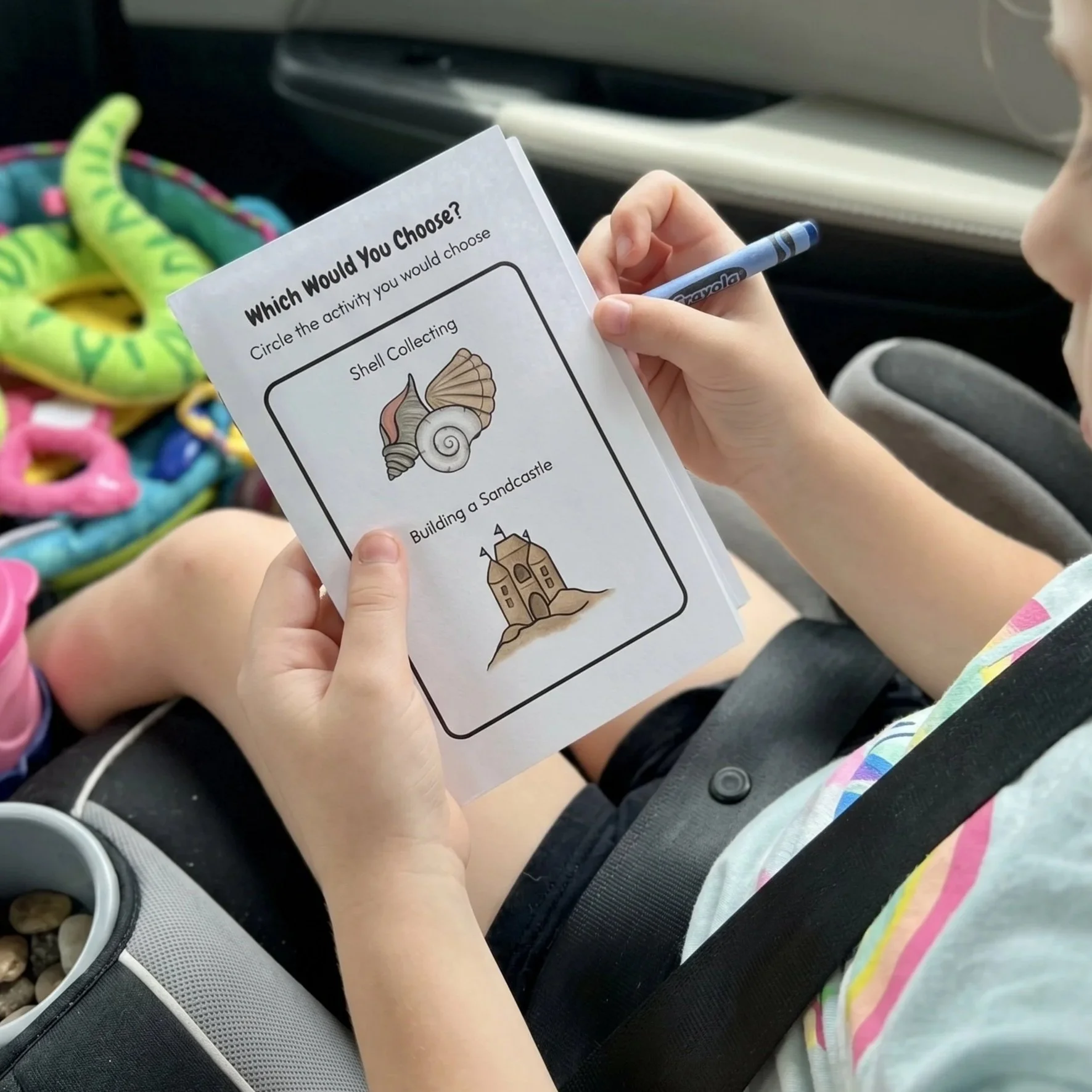 Child holding a sheet of paper with activity options, including shell collecting and building a sandcastle, in a car seat with colorful toys nearby.