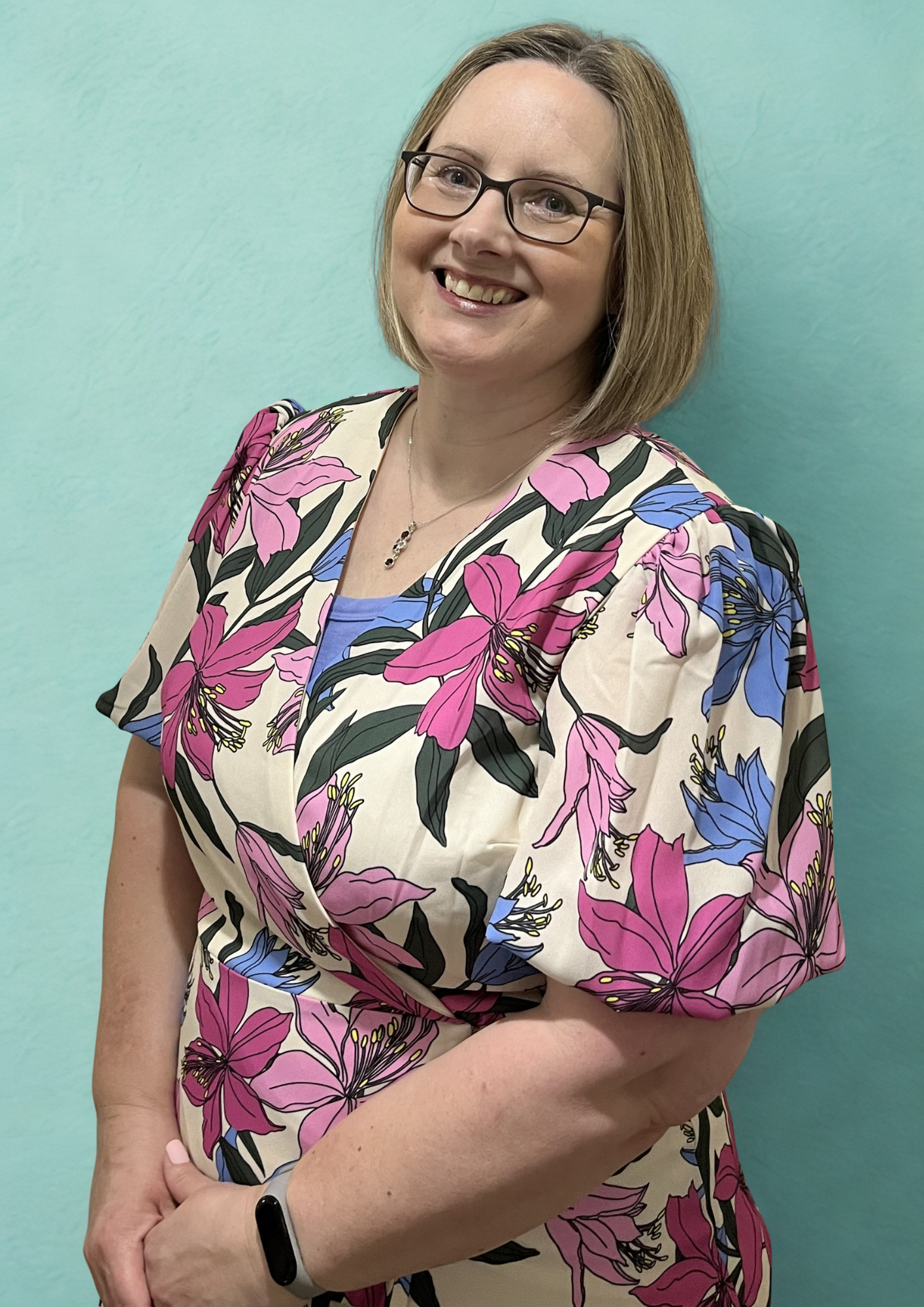 A woman with shoulder-length blonde hair, glasses, and a floral dress standing against a light green wall, smiling at the camera.