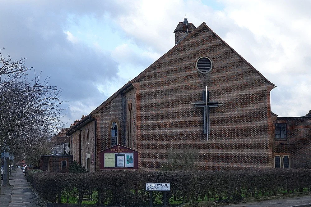 A brick church with a cross on the front wall and a small circular window near the roof. There is a signboard in front of the church and a street sign that reads 'Greenwood Road'.