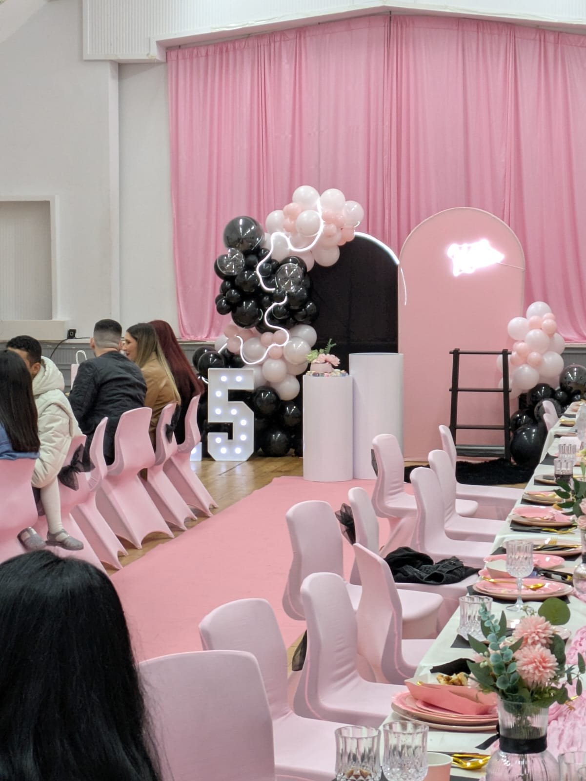 Event decorated with pink, black, and white balloons, a large illuminated number 5, pink drapes, and tables with pink and floral centerpieces celebrating a 5th birthday.