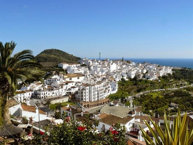 A scenic view of a white hillside town with numerous white buildings, green trees, and distant ocean under a clear blue sky.