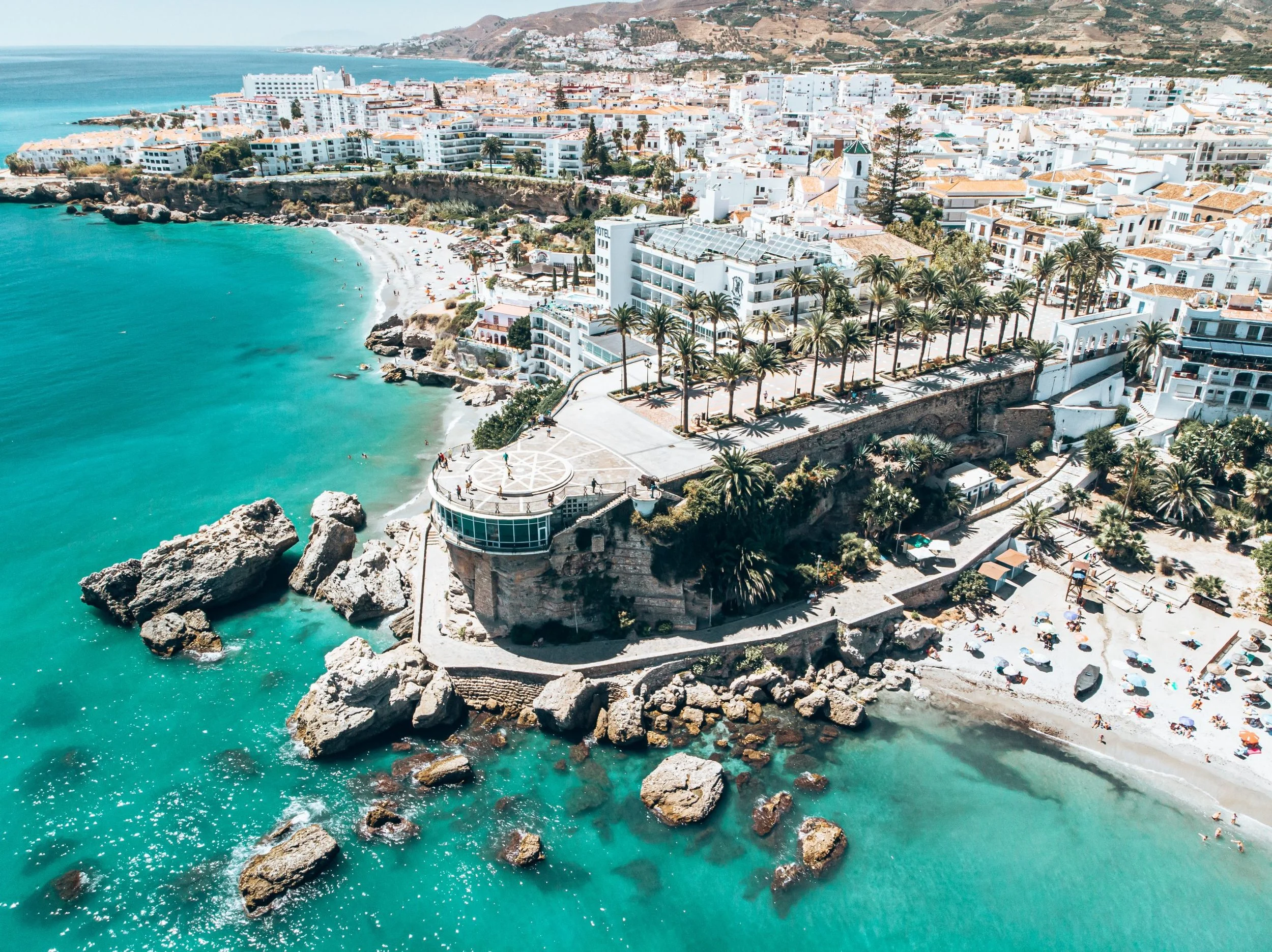 Aerial view of a coastal city with white buildings, a beach with umbrellas, palm trees, and turquoise ocean waters.