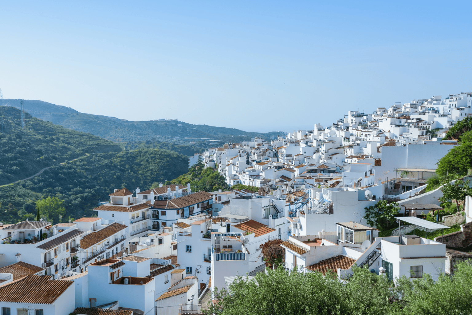 Hilly landscape with densely packed white buildings and terracotta roofs, surrounded by green hills and trees, under a clear blue sky.