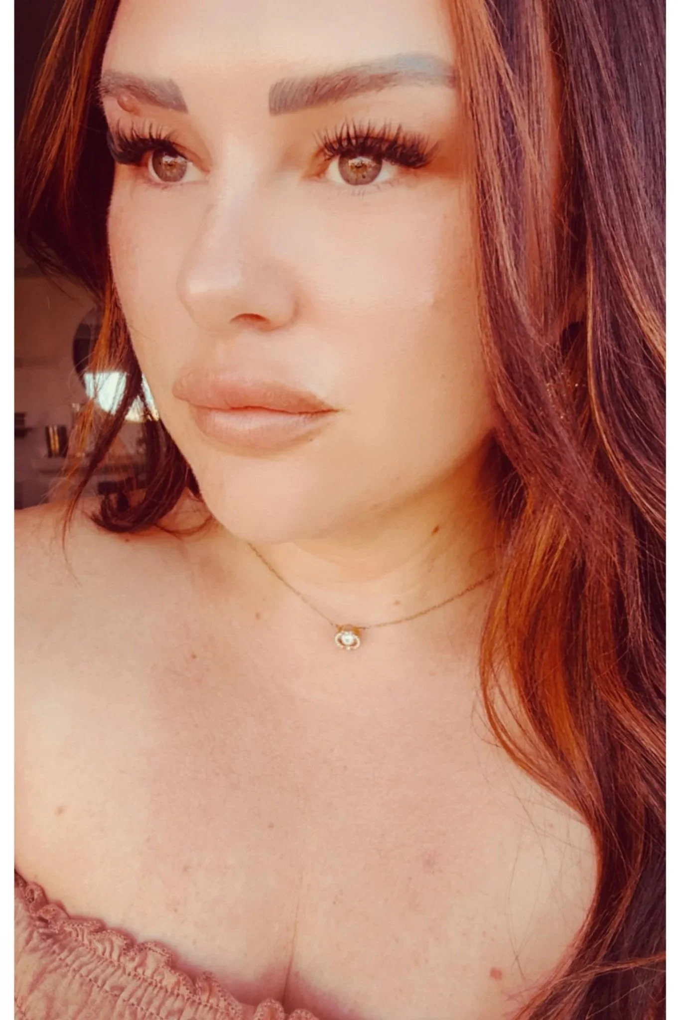 Close-up of a woman's face with brown eyes, long eyelashes, and arched eyebrows. She has long, wavy red hair and is wearing a delicate gold necklace with a small, round pendant. She is also dressed in a pink lace top.