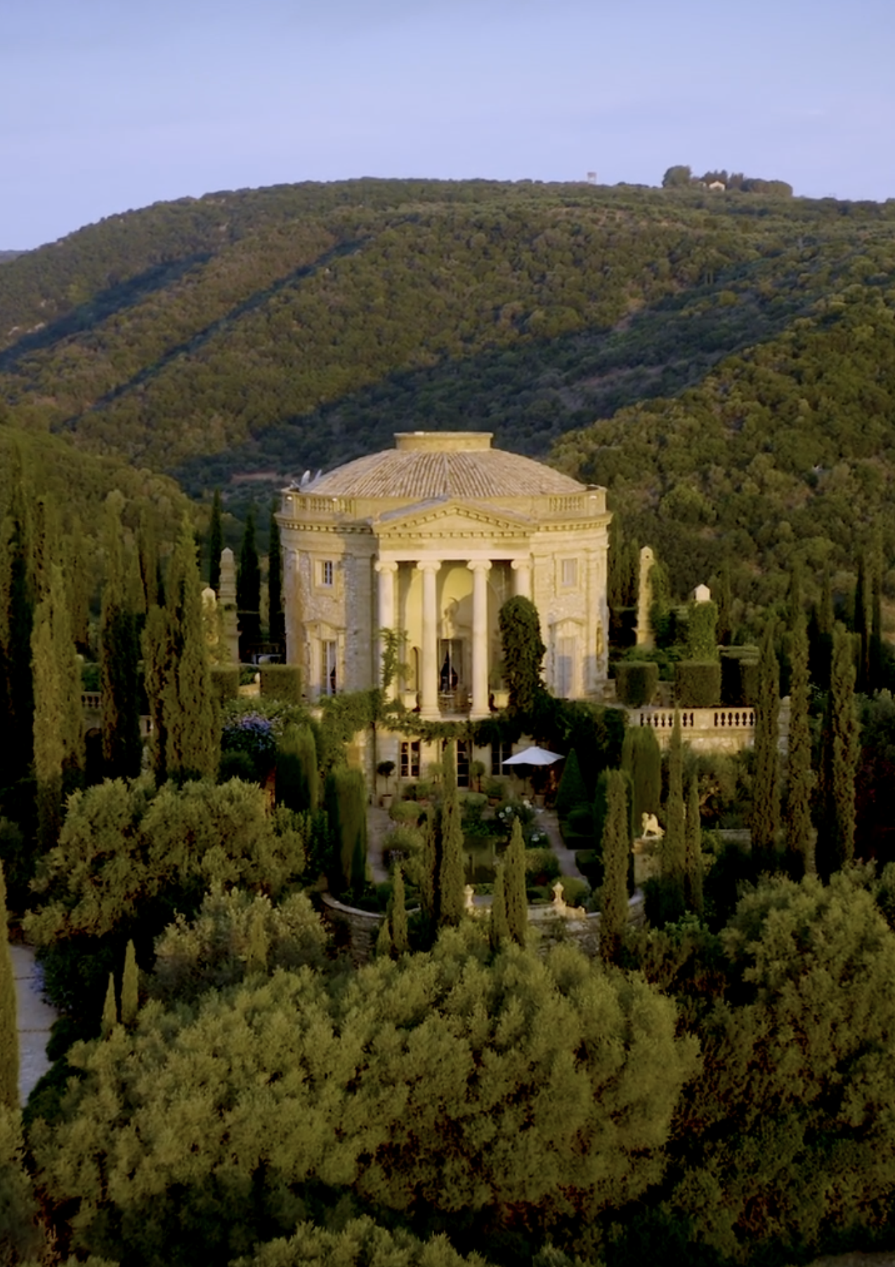 A large, circular building with a domed roof surrounded by lush green trees and cypress trees in a garden setting.