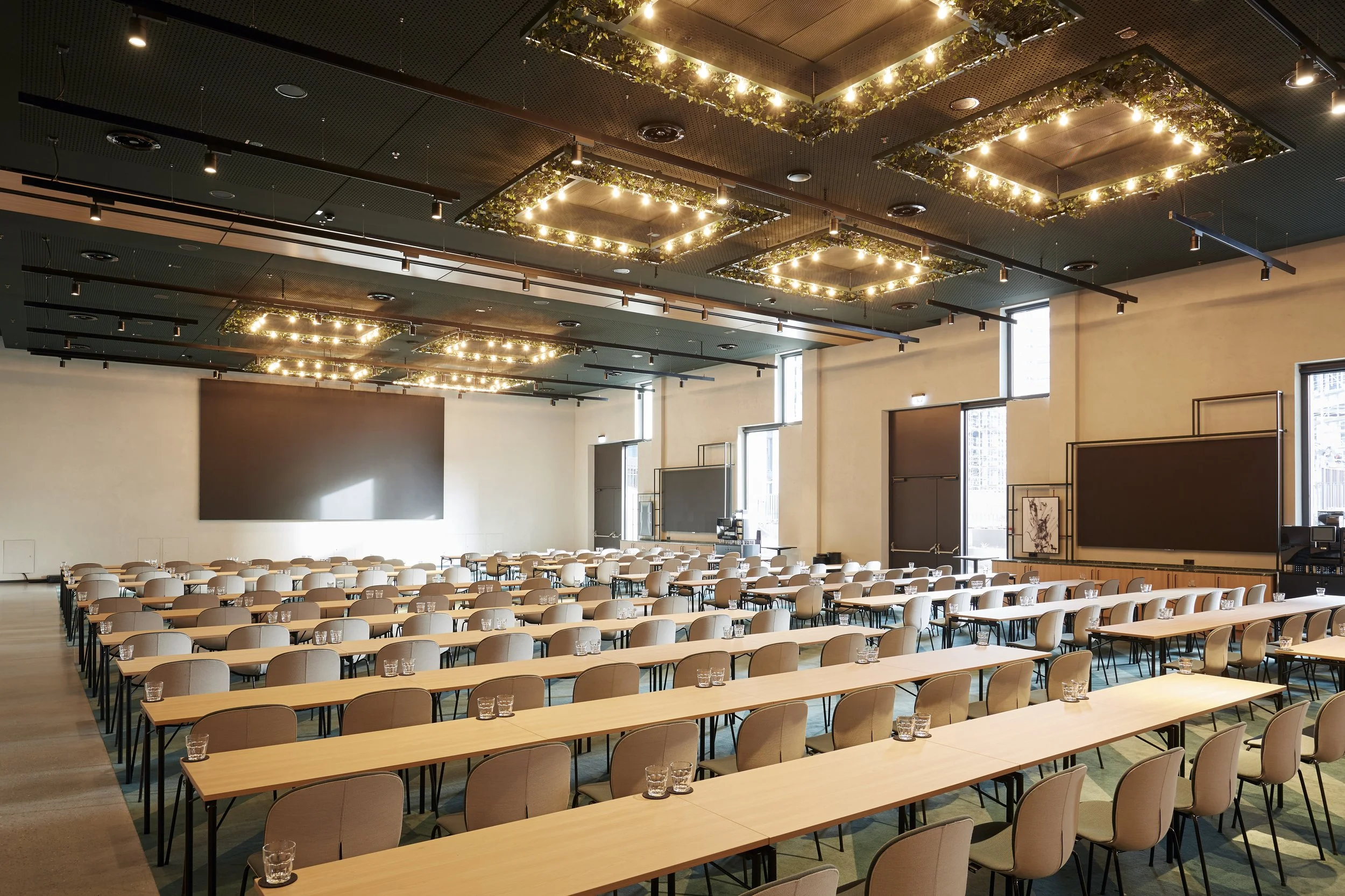 Ein großer, heller Versammlungsraum mit hohen Fenstern, vielen Stühlen und Tischen, dekorativen Beleuchtungen an der Decke und einer großen leeren Leinwand an der Wand.