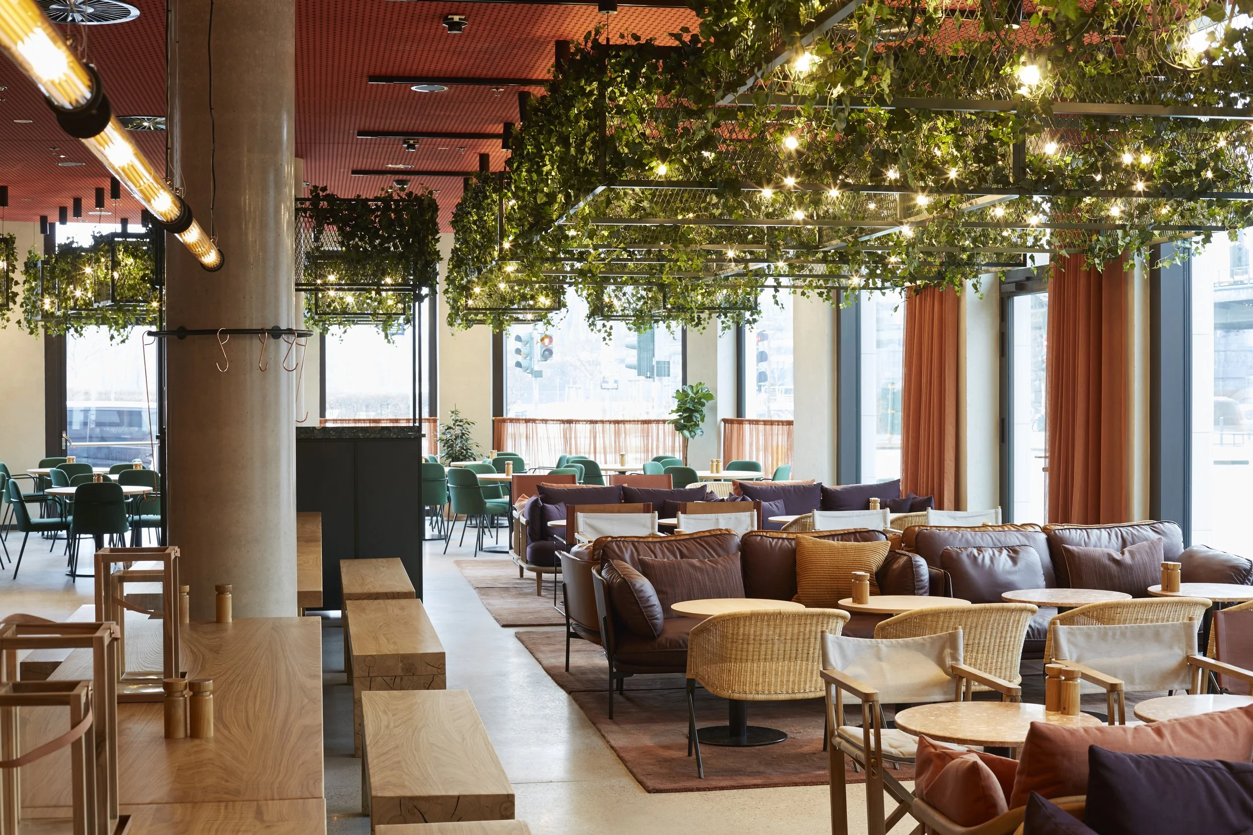 Innenansicht eines modernen Restaurants oder Cafés mit gemütlicher Einrichtung, grünen und beigen Sitzmöbeln, großen Fenstern, und einer Decke mit grünen Pflanzen und Lichterketten.