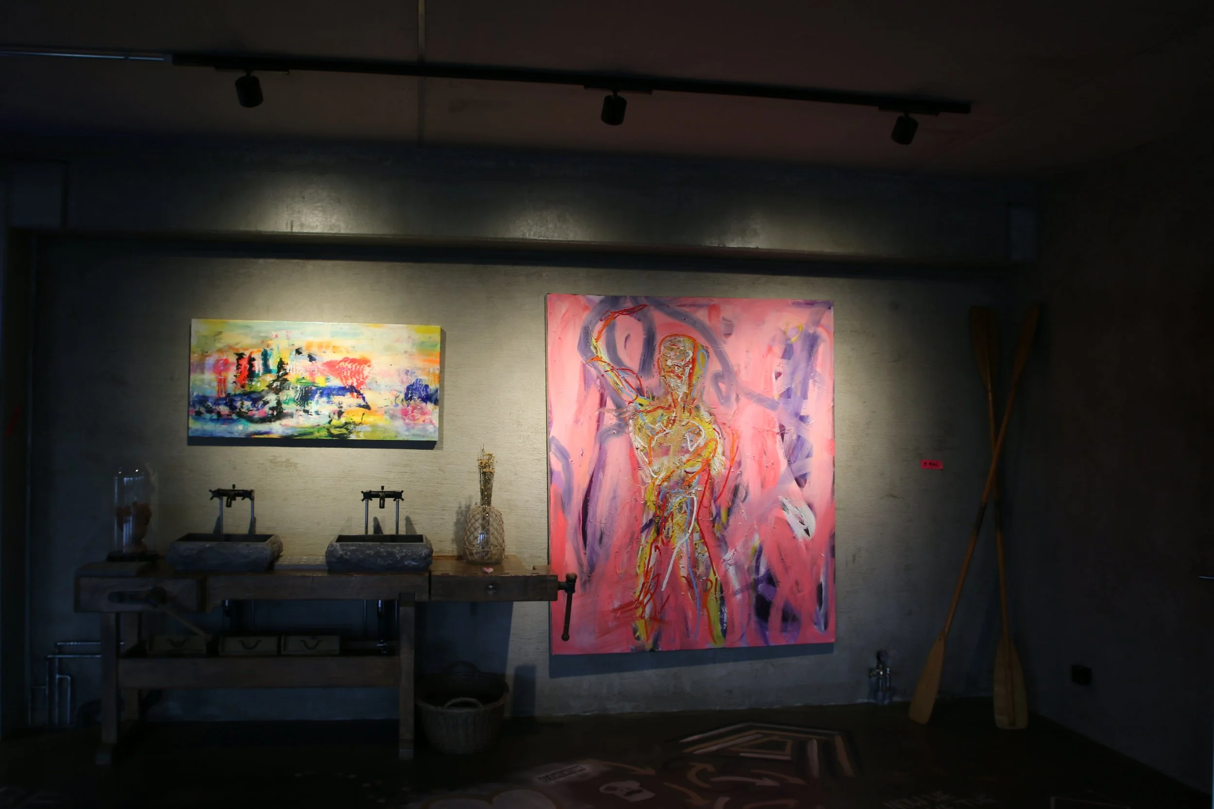 Gemälde in einer Galerie, ein großes Bild mit abstrakten, lebendigen Farben, neben einem weiteren abstrakten Kunstwerk, darunter eine Werkbank mit Malutensilien.