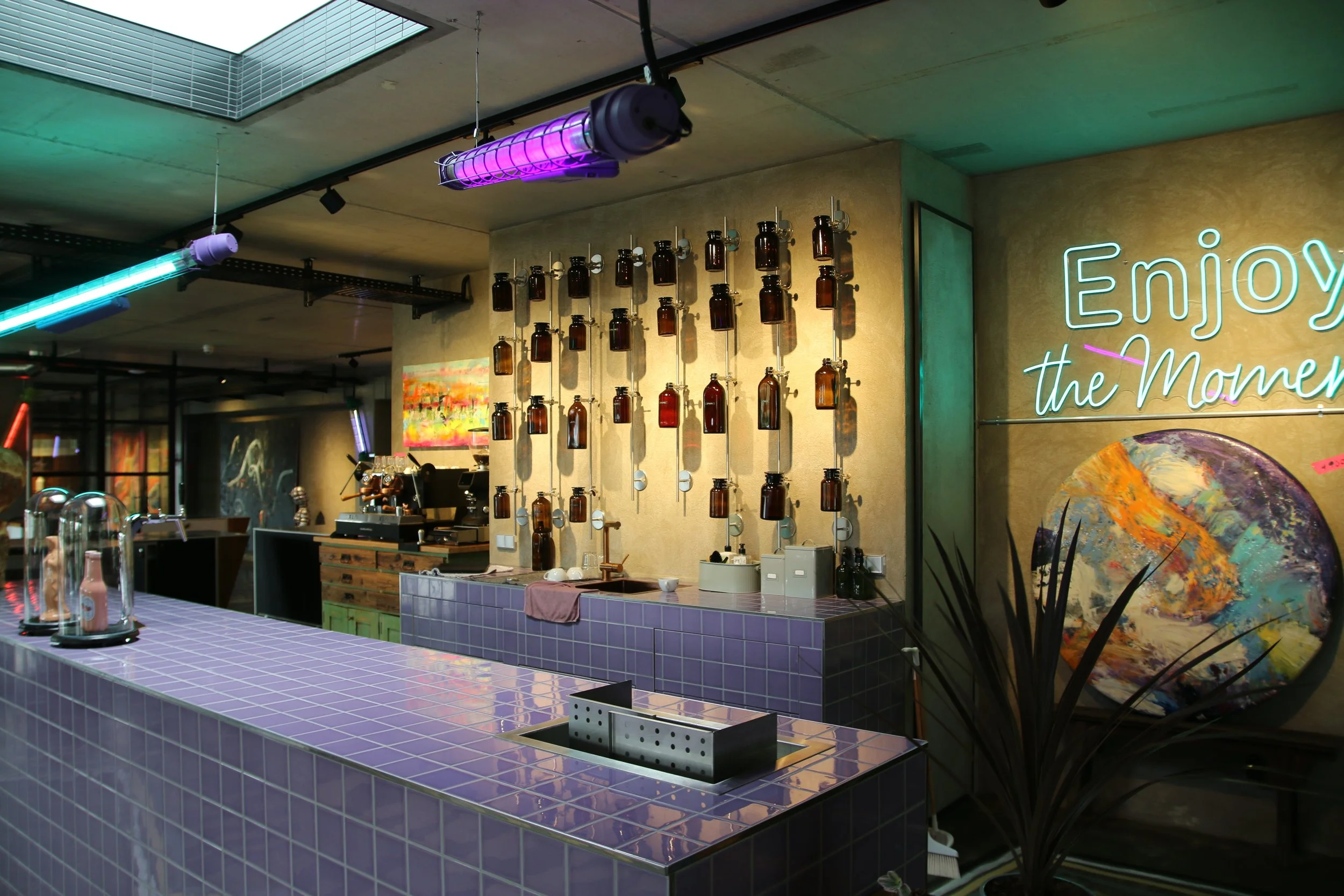 Ein moderner Café- oder Barbereich mit einer Theke aus violett-glasierten Fliesen, dekorativen Flaschen an der Wand, Neon-Schriftzug 'Enjoy the Moment' und farbigen Lichtakzenten an der Decke.