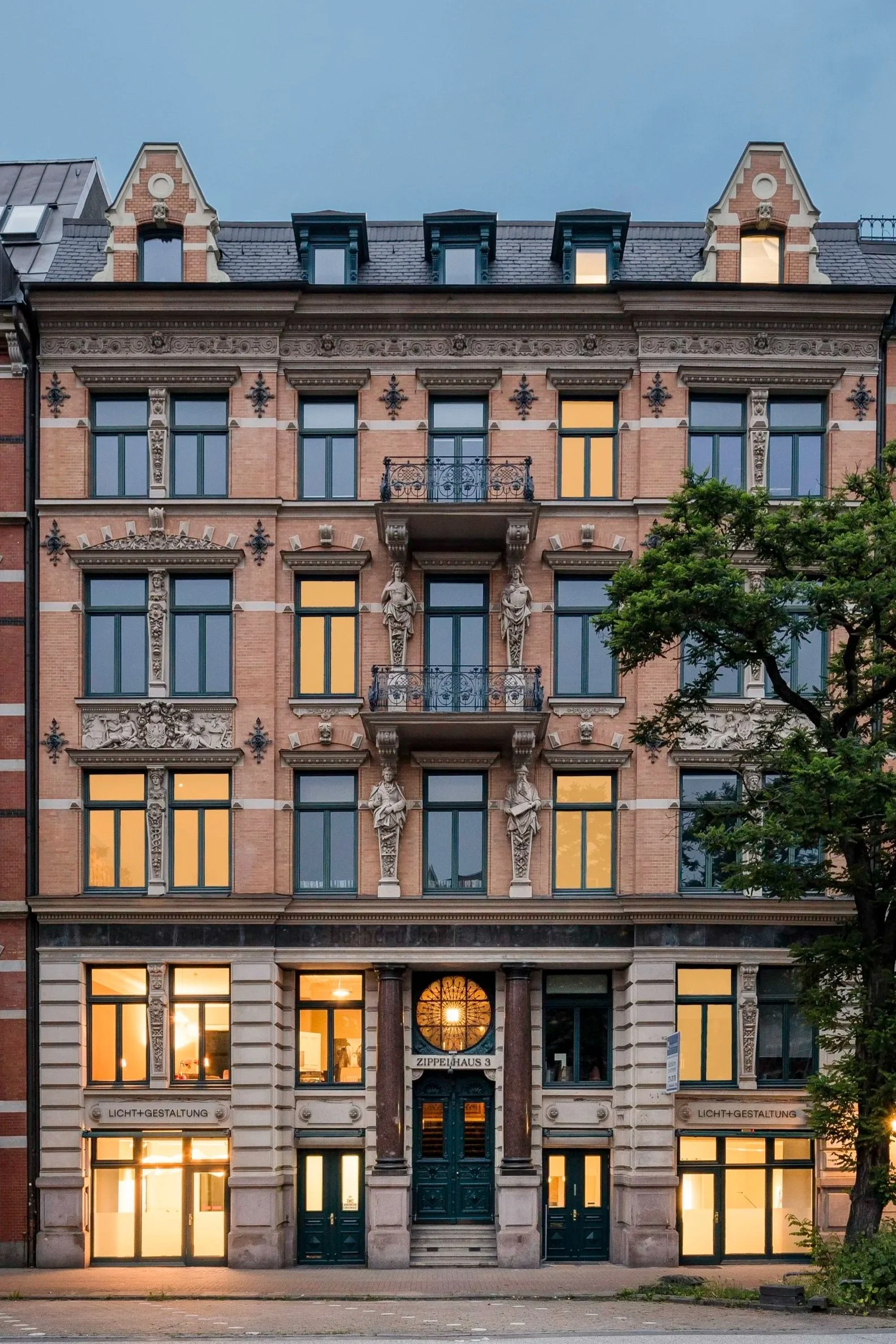 Historisches Gebäude mit barocker Fassade, Statuen an der Fassade, Balkon mit schmiedeeisernem Geländer, große Fenster mit warmem Licht, Baum links im Vorfeld, Abendstimmung
