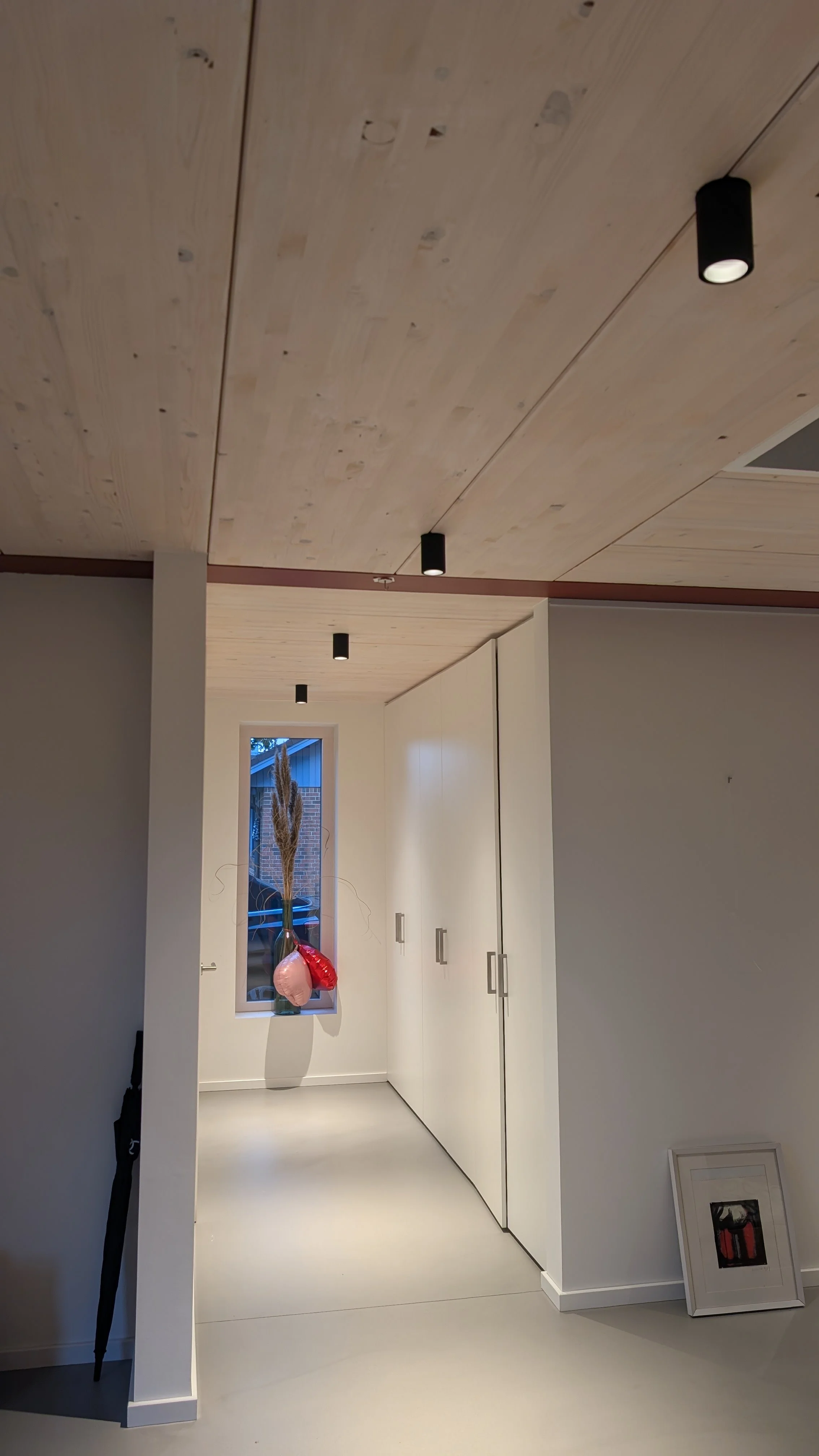 Innenansicht eines Raumes mit hellen Wänden und weißen Schränken, eine Fenster mit Sicht nach draußen, Dekoration mit Ballons und ein Bild im Rahmen auf dem Boden, eine Decke mit schwarzen Spotlights.