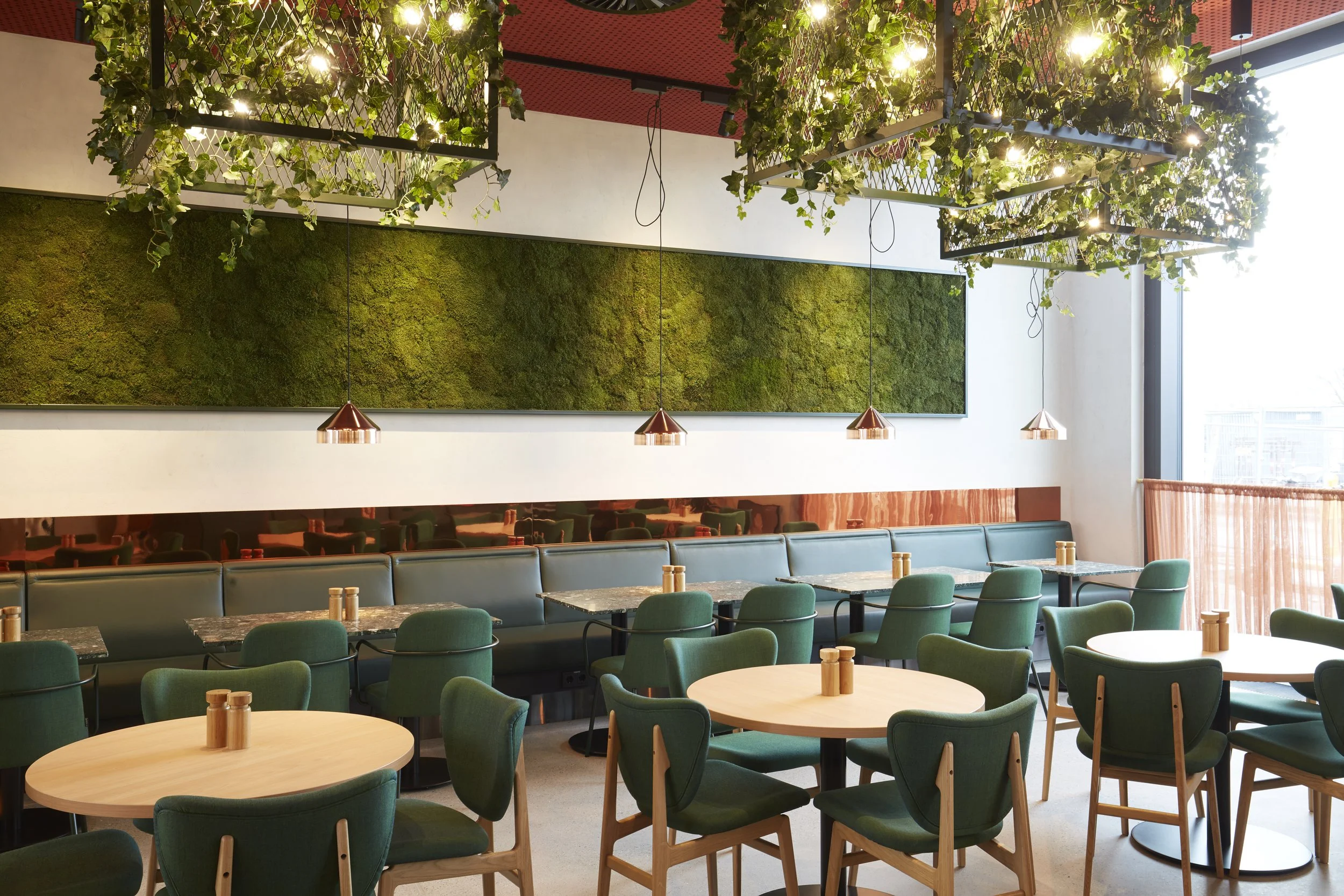 Innenraum eines modernen Restaurants mit grünen Stühlen, runden Tischen, einer begrünten Wand mit Moos, hängenden Kupferlampen und viel natürlichem Licht durch große Fenster.