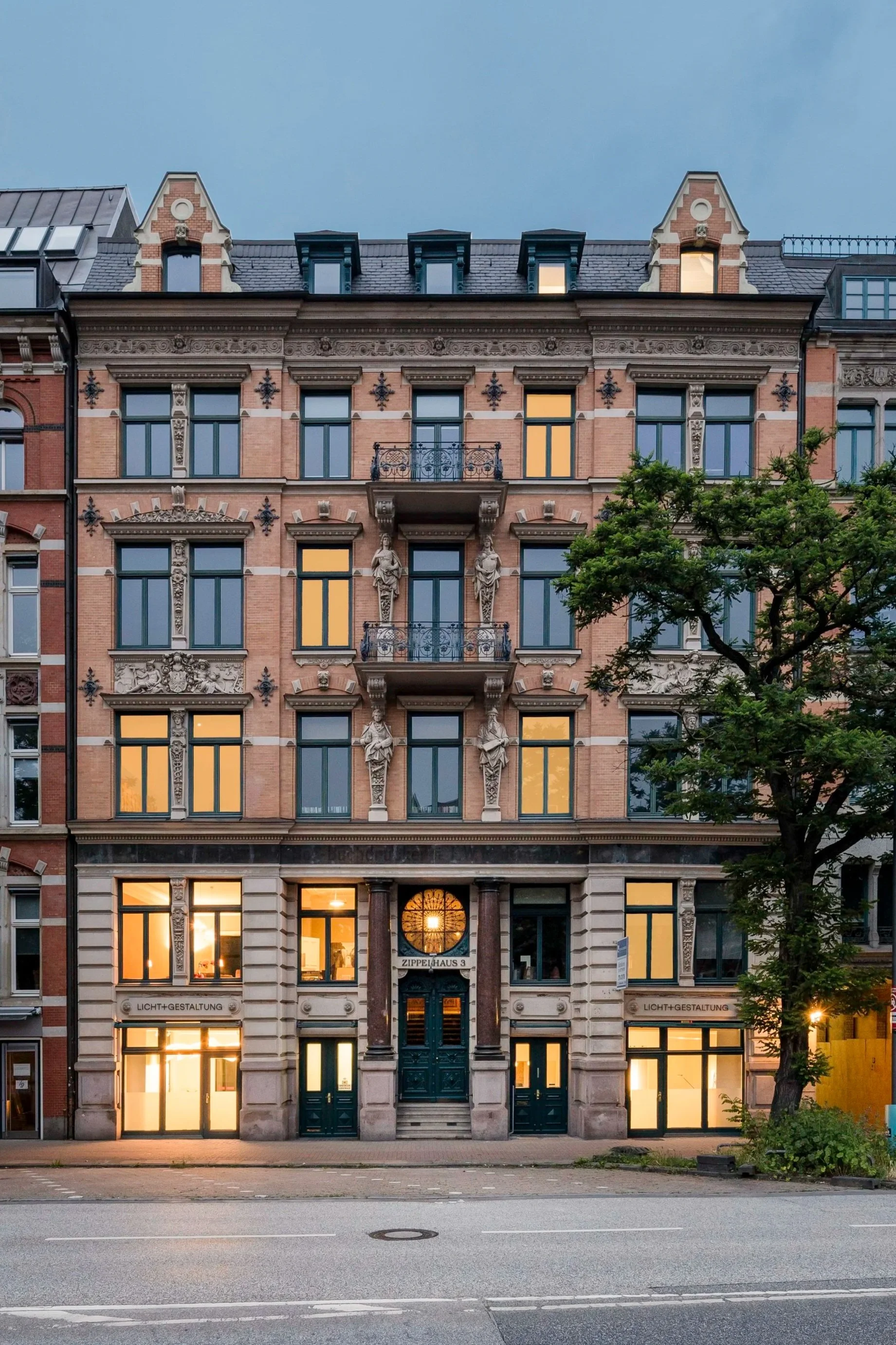 Mehrstöckiges historisches Gebäude mit ornamentalen Fassadendetails, Figuren und Balkonen, beleuchtet bei Sonnenuntergang, mit Baum im Vordergrund.