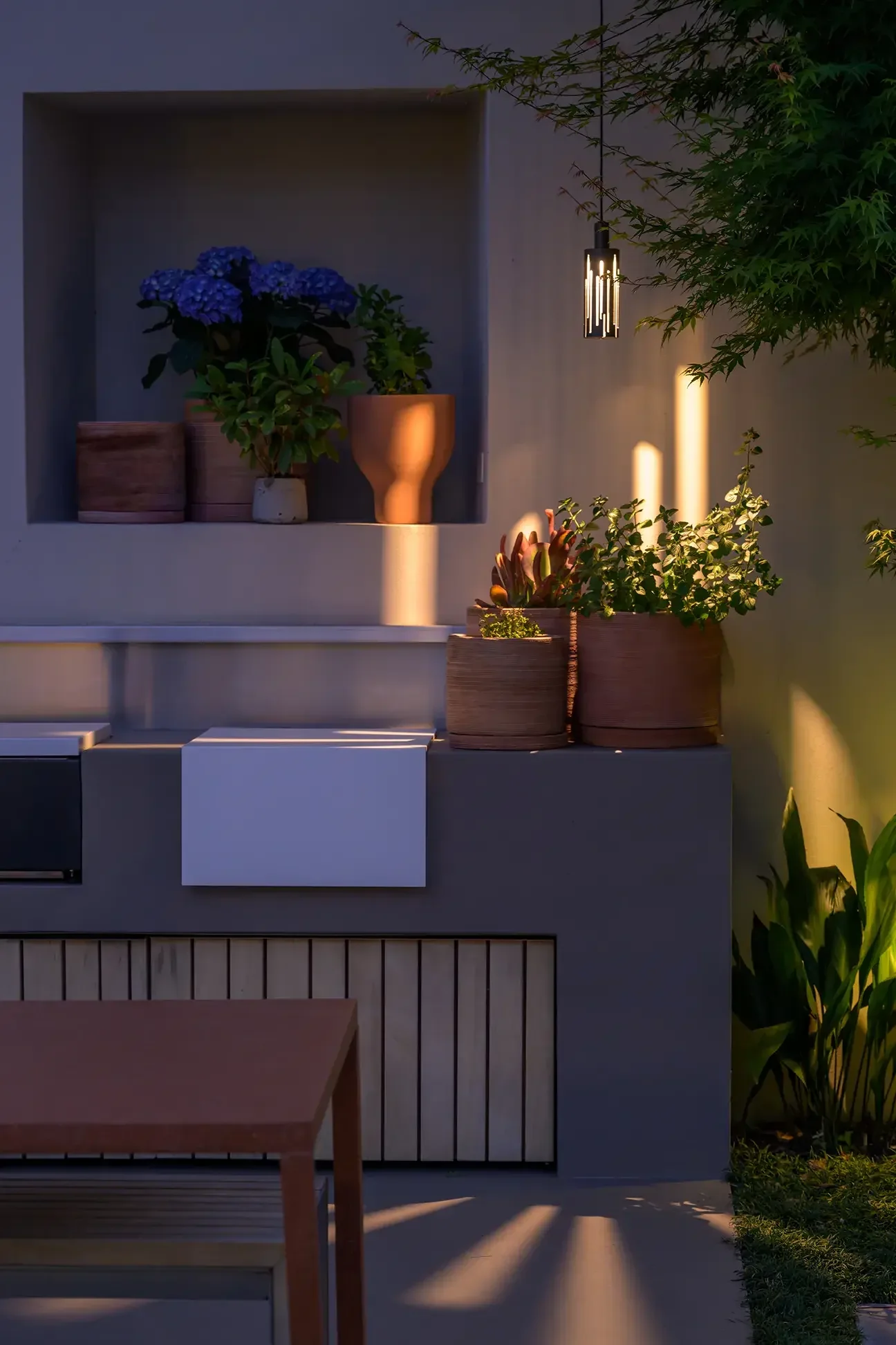 Blick auf eine moderne Terrasse mit gedecktem Bereich, Blumentöpfen und einem hölzernen Tisch, bei Sonnenuntergang