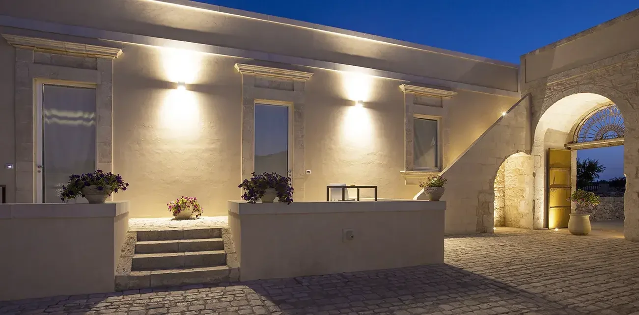 Ein abendlicher Blick auf eine weiß getünchte, historische Fassade mit beleuchteten Fenstern und Blumenvasen davor, einschließlich eines Bogens und einer offenen Tür, auf einer gepflasterten Terrasse.