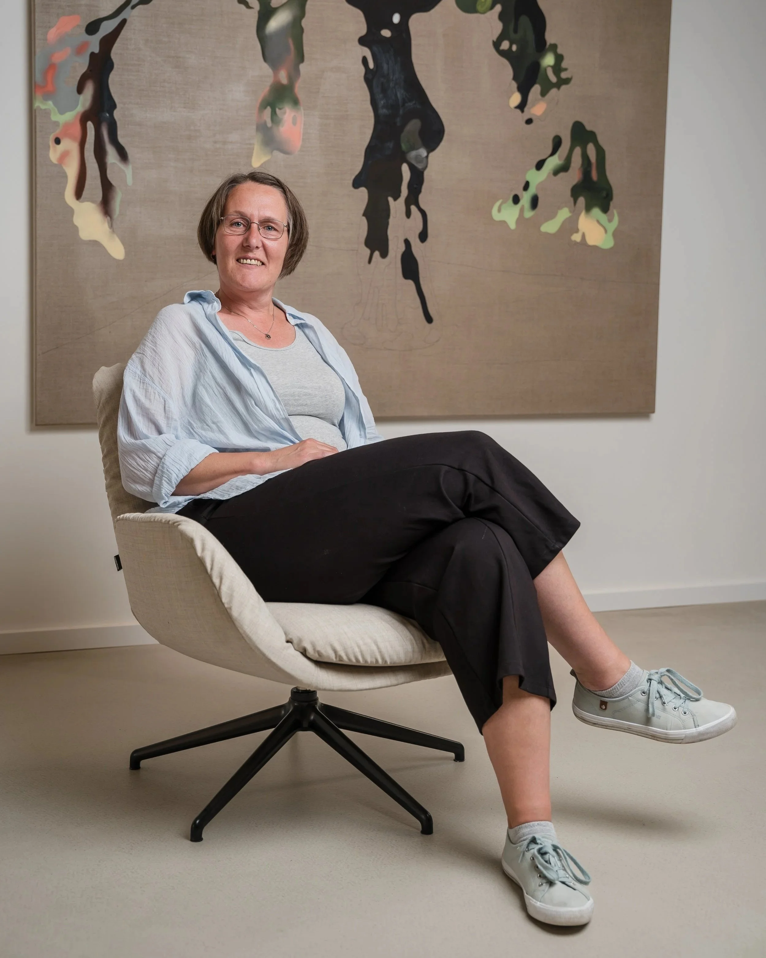Eine Frau sitzt entspannt in einem modernen weißen Bürostuhl vor einer abstrakten Wandkunst in neutralen Farben, die aus schwarzem, grünem, pinkem und beige Farben besteht. Sie trägt eine graue T-Shirt, eine offen helle Bluse, schwarze Hosen, und weiße Sneakers. Ihre kurzen braunen Haare sind leicht gewellt, und sie trägt eine Brille. Die Wand hinter ihr ist hell und die Atmosphäre wirkt modern und komfortabel.