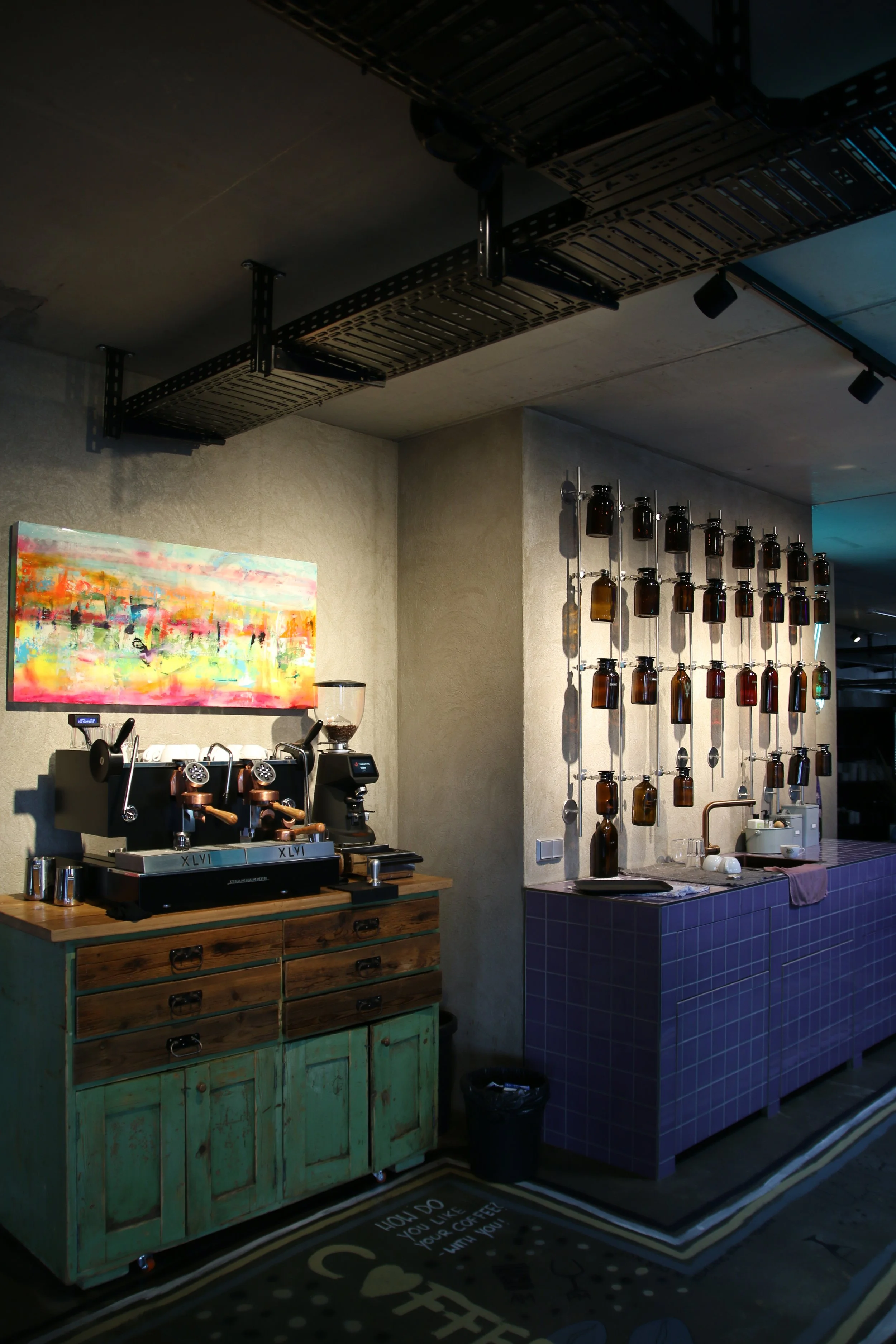 Eine Café- oder Bar-Umgebung mit einem Kaffeemaschinen-Bereich, einem bunte bemalten Holzschrank, einer Wand mit vielen braunen Flaschen und einem blauen, gefliesten Bartresen. Die Wand ist aus hellem Beton, und es gibt einen bunten abstrahierten Kun