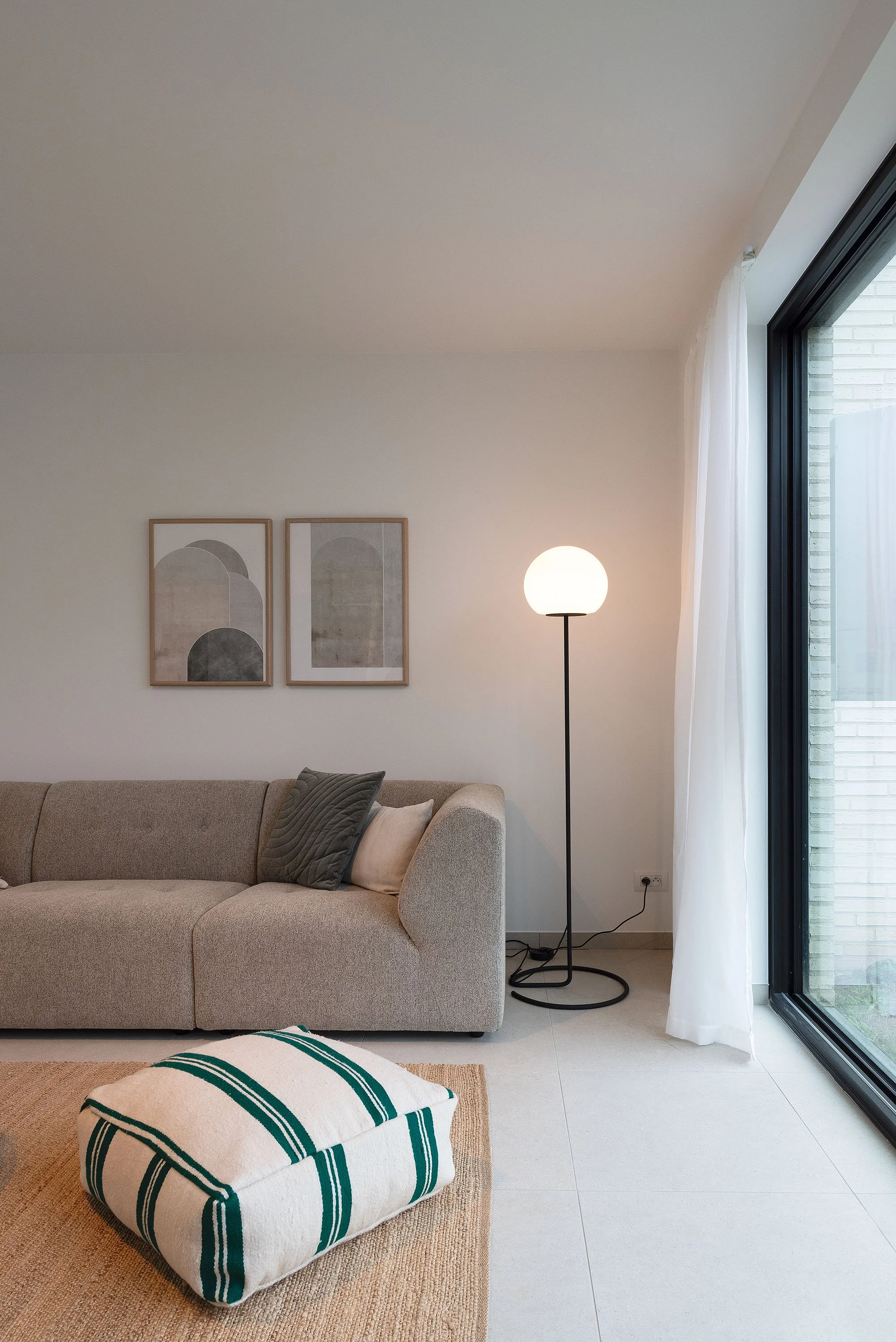 Ein Wohnzimmer mit einem beige Sofastück, einem Kissen, einem weißen und grünen gepolsterten Hocker auf einem braunen Teppich, einer Stehlampe mit kugelförmigem Lampenschirm, zwei Wandbilder mit geometrischen Mustern, großen Fenstern mit weißen Vorhä