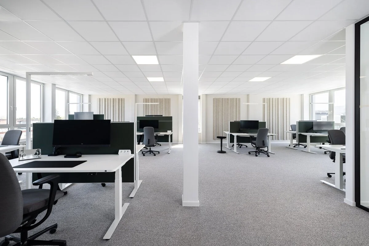 Leeres modernes Büro mit weißen Tischen, schwarzen Bildschirmen und Bürostühlen, große Fenster, grauer Teppichboden, weiße Wände, Decken mit quadratischen Paneelen.
