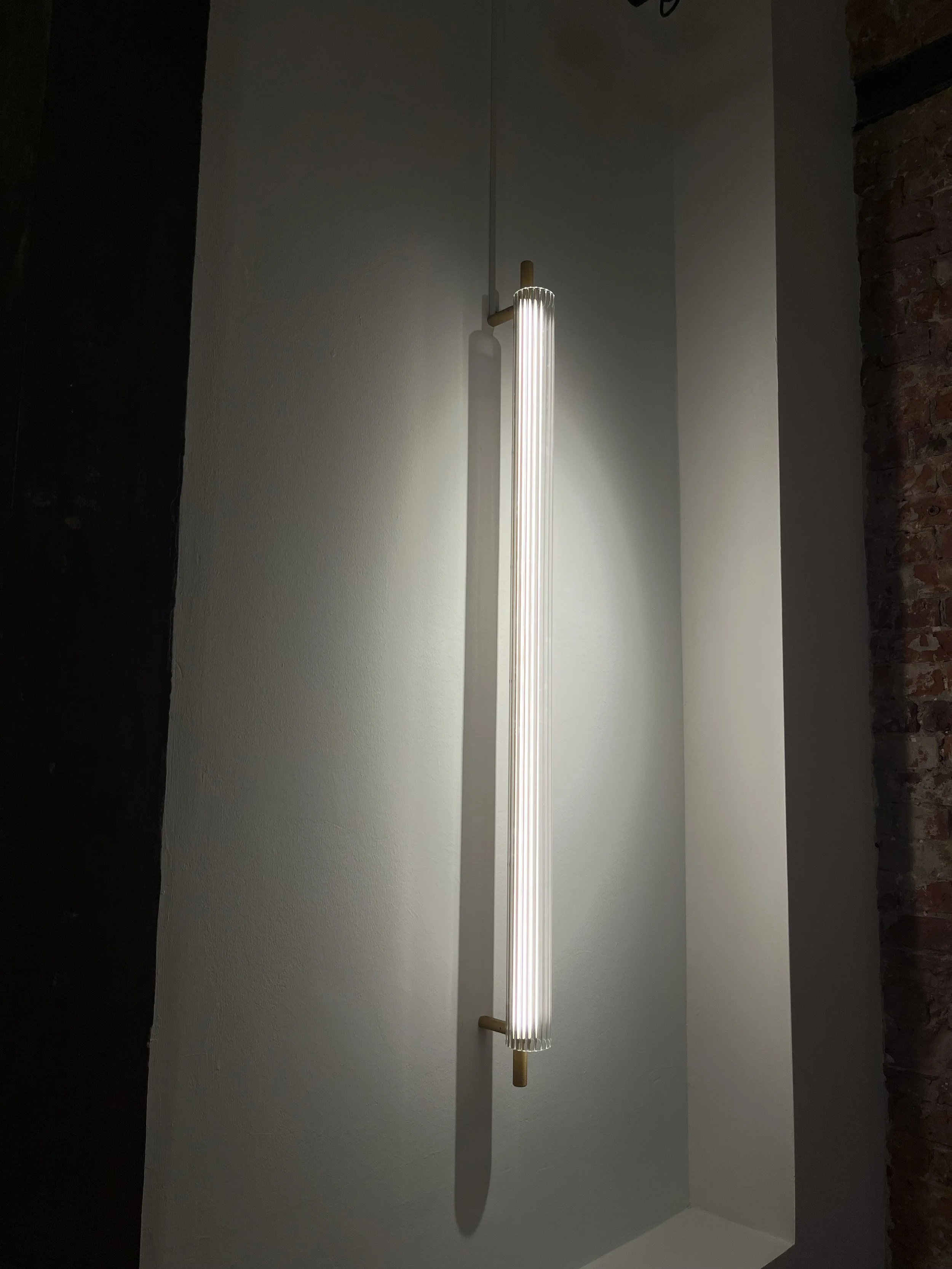 Moderne Wandlampe mit goldfarbenem Arm und weißem Leuchtstab, an einer weißen Wand in einem Raum mit dunklen Ziegelwänden.