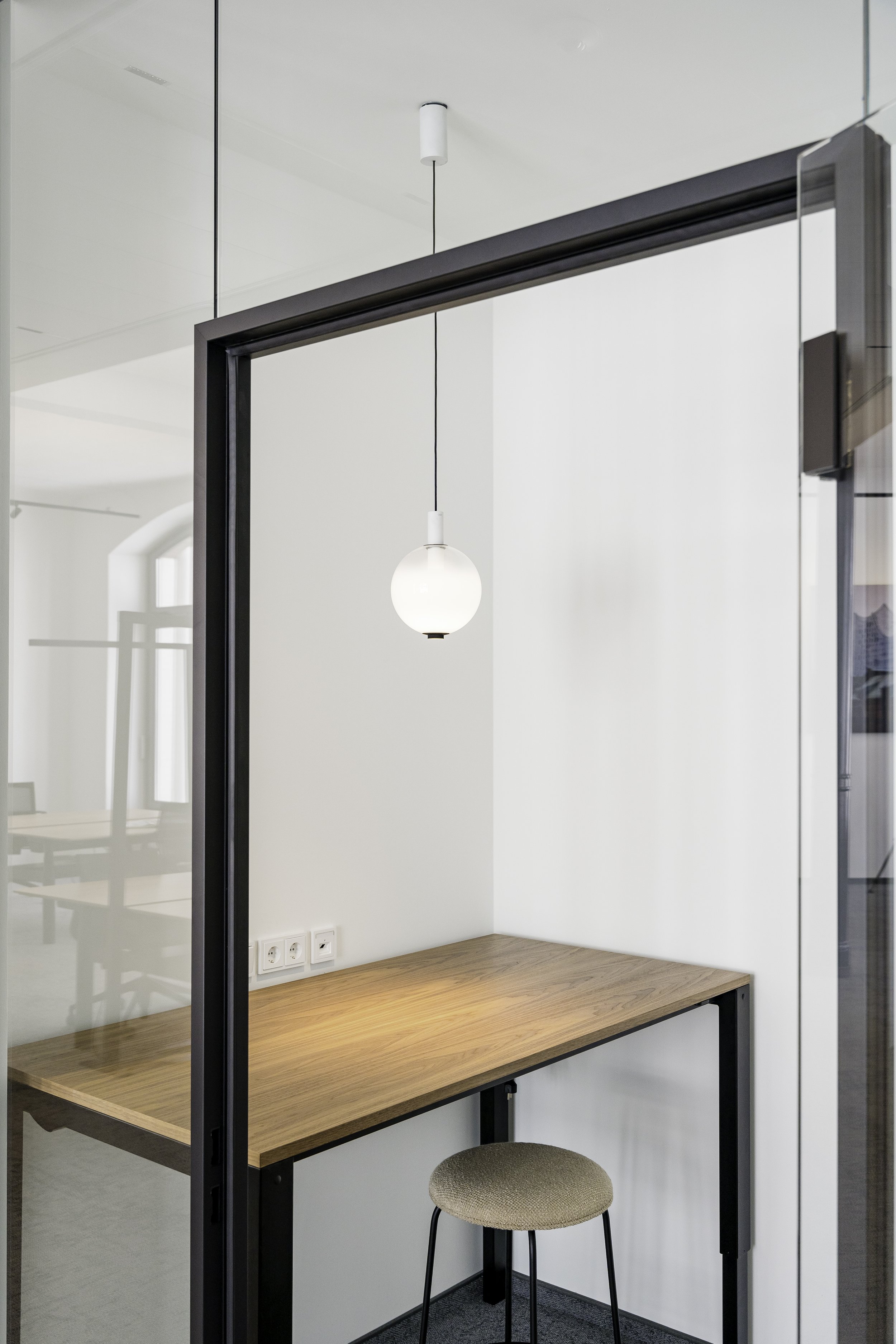 Ein moderner Arbeitsplatz mit einem Holzschreibtisch, einem runden Hocker und einer weißen Pendant-Lampe, in einem Bereich mit weißen Wänden und mehreren Stromsteckdosen.