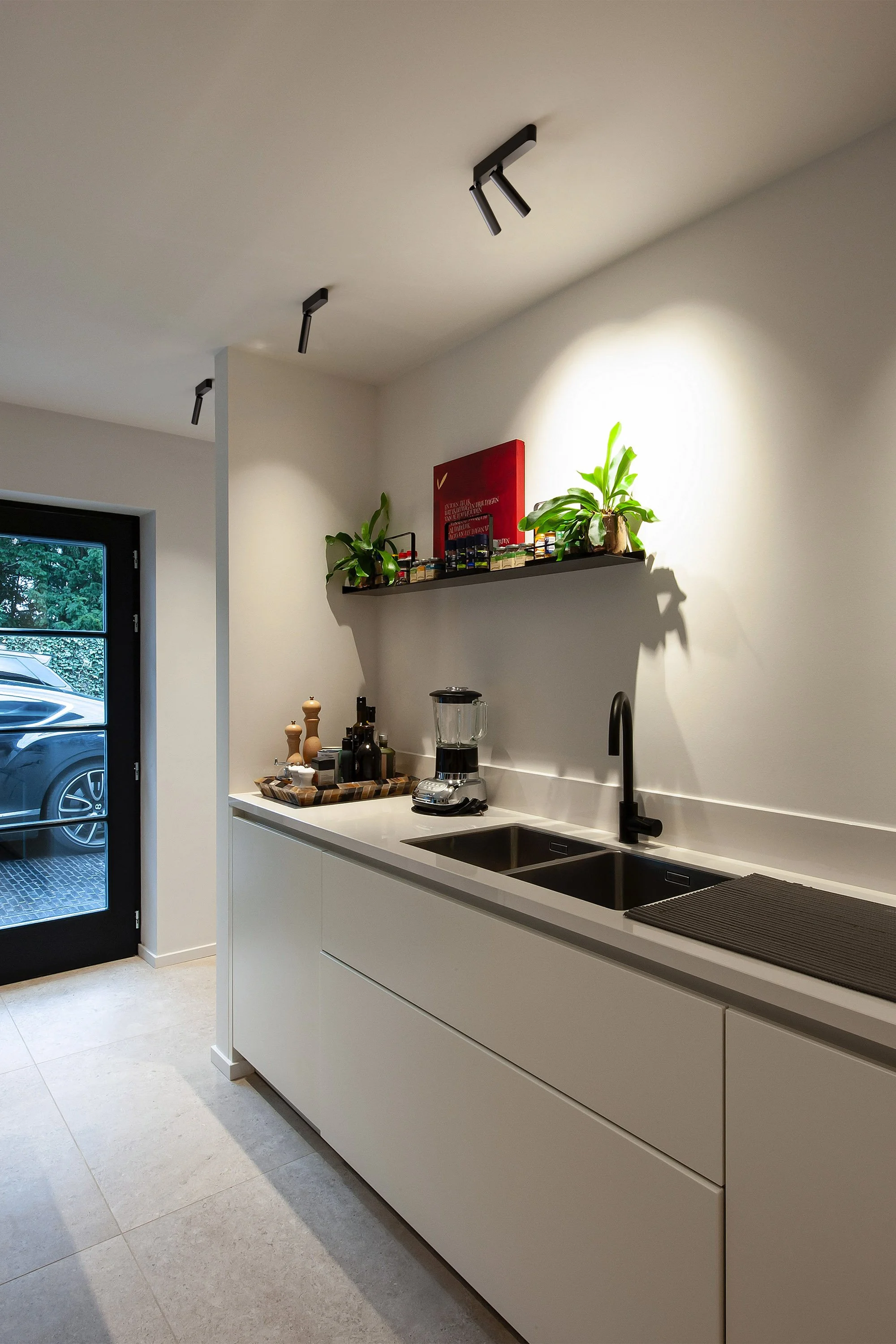 Moderne Küche mit weißem Korpus, schwarzen Armaturen, zwei Becken, auf der Ablage stehen Pflanzen und Küchenutensilien, es gibt einen Herd und eine Kaffeemaschine, an der Wand hängt ein rotes Bild.