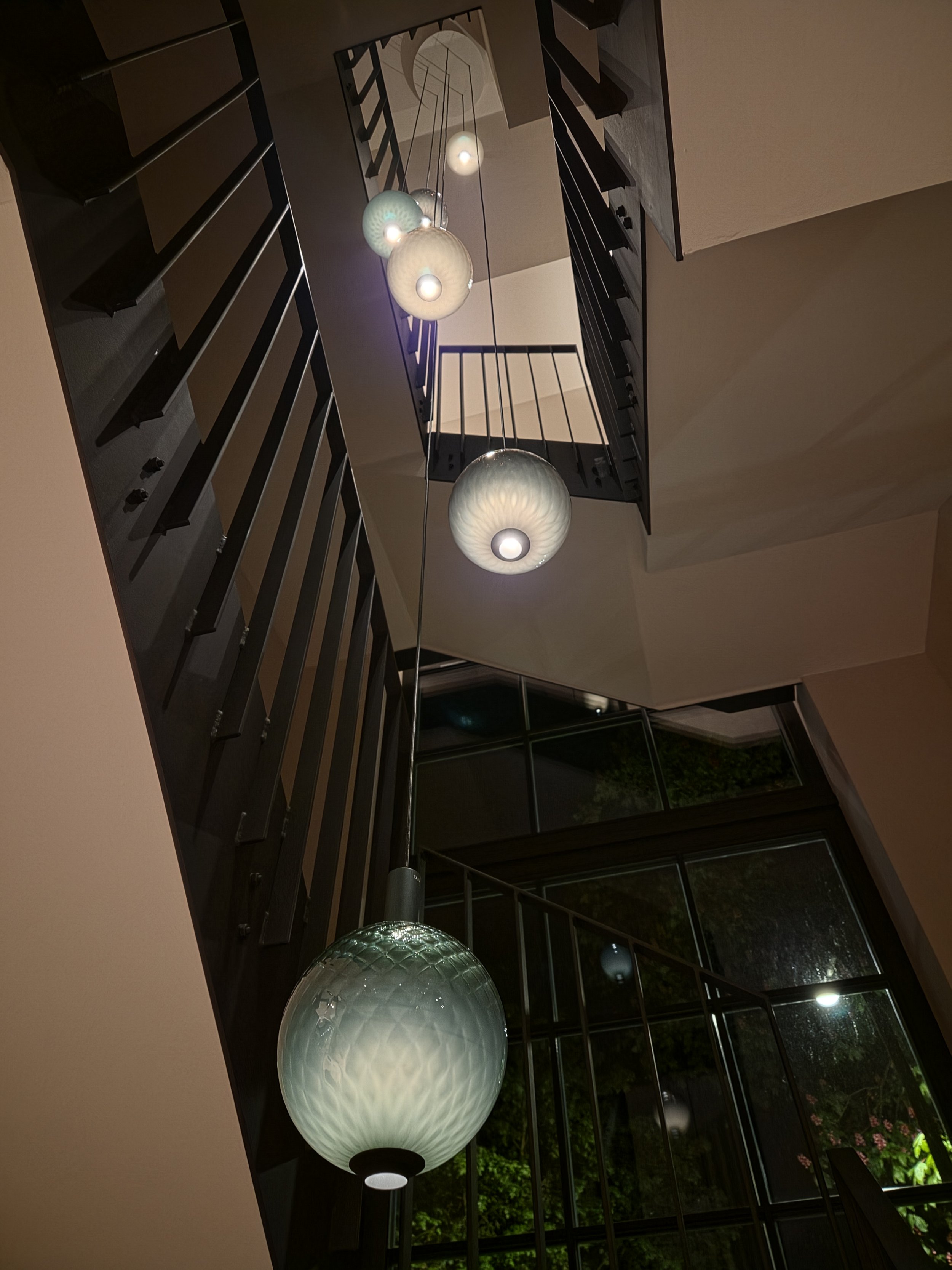 Lampen hängen von der Decke eines Treppenhauses, Blick nach oben, mit Fenstern im Hintergrund und Bäumen draußen.