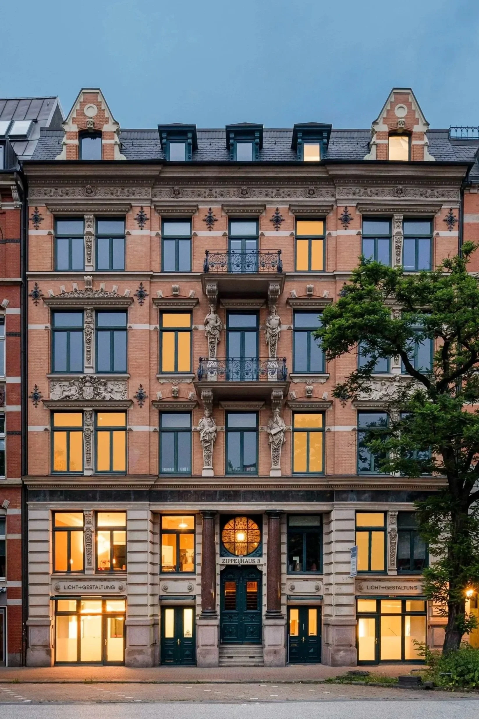 Historisches Gebäude mit kunstvollen Fassadendekorationen, mehreren Fenstern und einem Eingang, beleuchtet im Abendlicht.