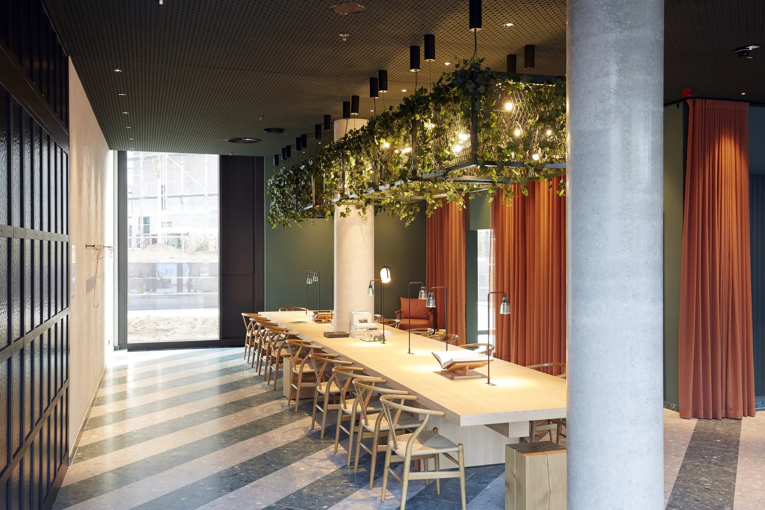 Innenansicht eines modernen Restaurants mit langer Holztheke, orange Vorhängen, grüner Wand, großen Fenstern, und einer dekorativen Deckenleuchte mit Pflanzen und Lichtern.