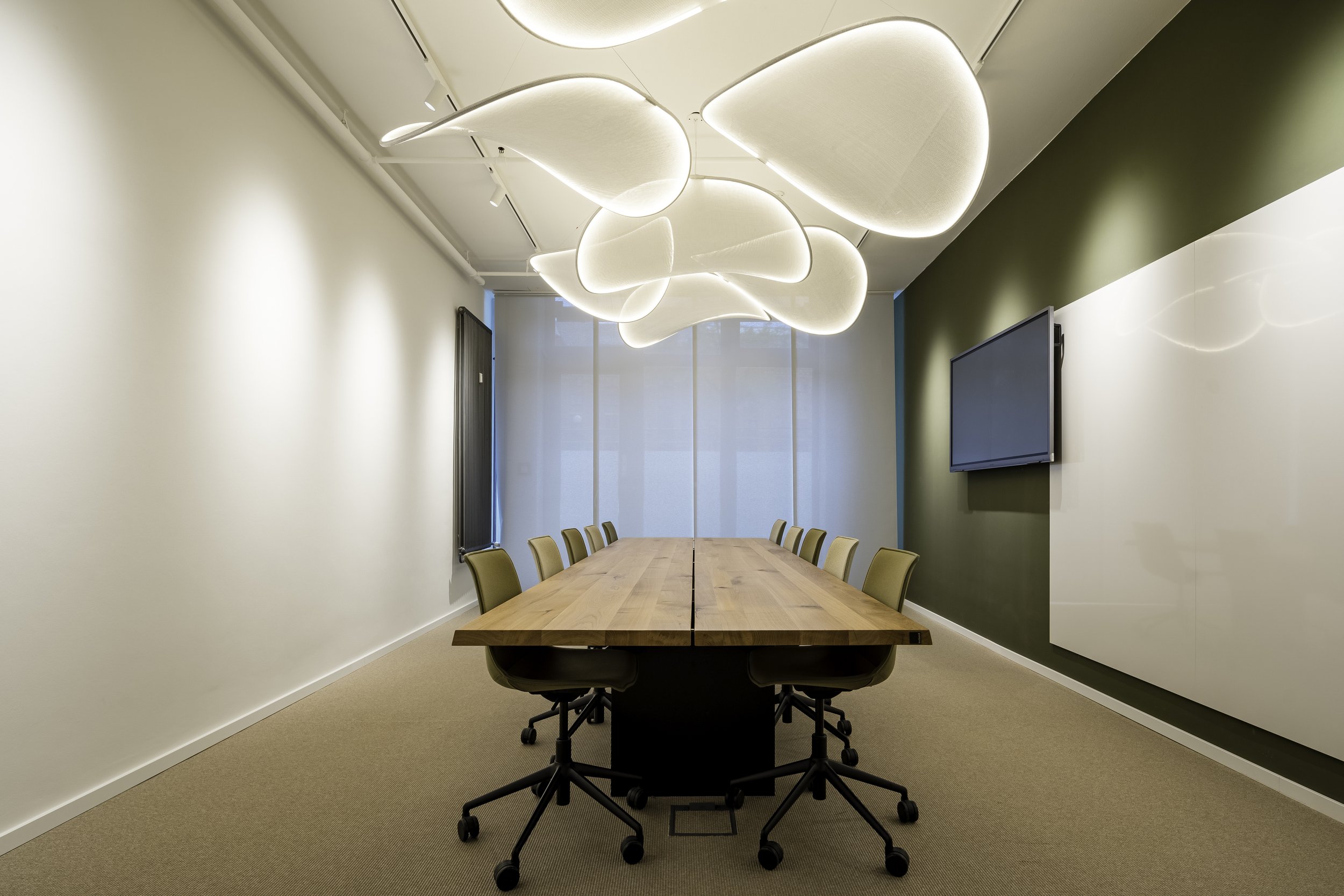 Ein moderner Konferenzraum mit einem langen Holztisch, umgeben von schwarzen Bürostühlen. An der Wand hängt ein großer Bildschirm. Über dem Tisch sind stylische weiße Deckenleuchten in organischer Form. Die Wände sind hell, eine Wand ist grün gestric