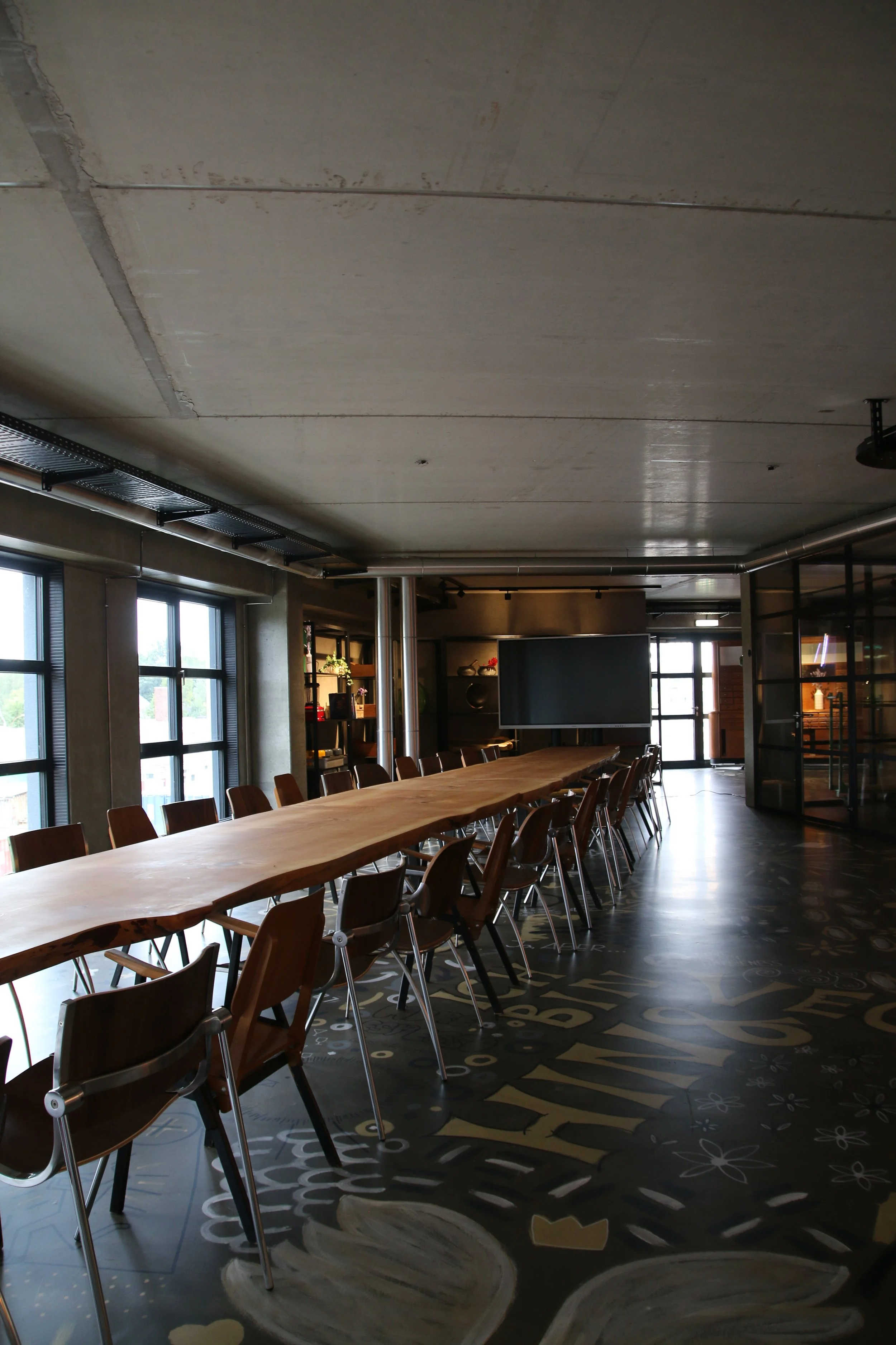 Ein moderner, heller Konferenzraum mit einem langen Holztisch, Stühlen und großen Fenstern, die Tageslicht spenden. Es gibt eine große Bildschirmanzeige an der Wand und Regale mit Dekorationsgegenständen. Der Boden hat ein Muster mit Blumen und geome