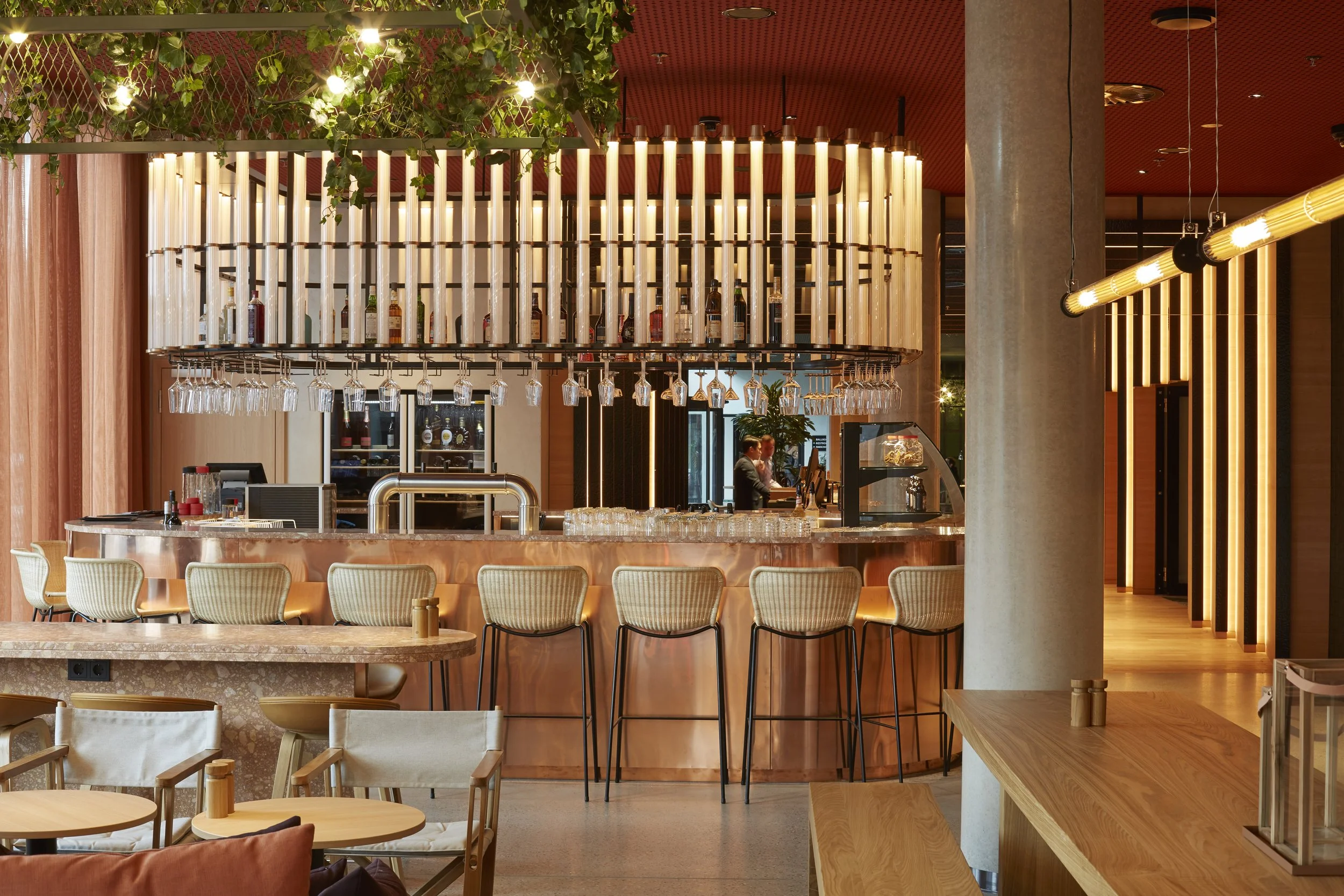 Innenansicht eines modernen Cafés mit Holz- und Kupfer-Akzenten, Bar mit Hängelampen, Sitzgelegenheiten, Getränke an der Bar, Pflanzen und warmes Licht.