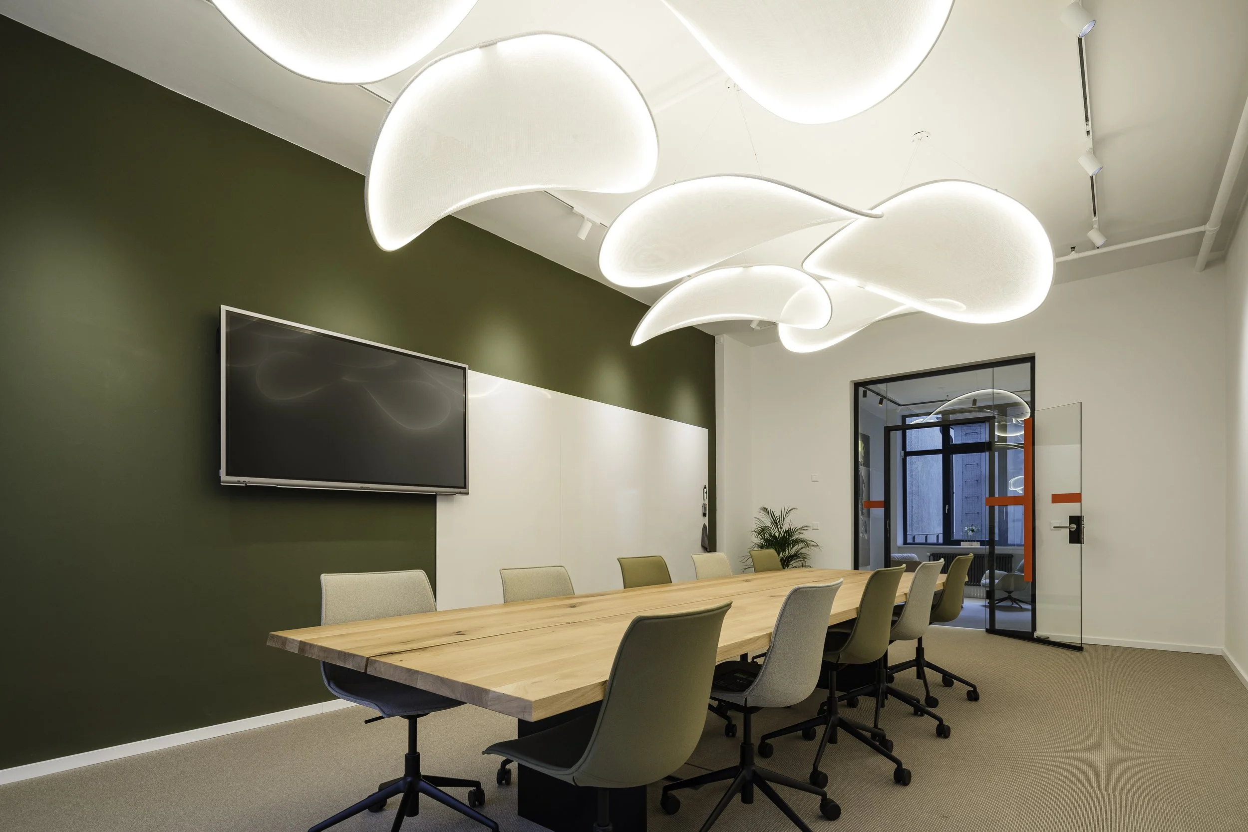 Ein modern eingerichteter Konferenzraum mit einem großen Holztisch und mehreren Stühlen. An der Wand hängt ein großer Bildschirm. Die Decke ist mit einer weißen, geschwungenen Lichtinstallation ausgestattet. Es gibt eine Tür mit einer Glastrennwand im Hintergrund.