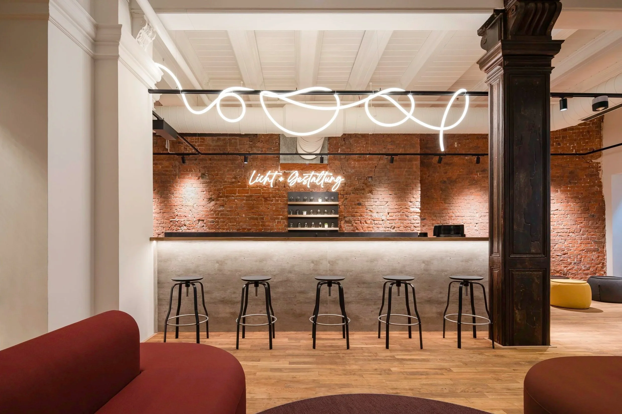 Innenansicht eines modernen Cafés mit Barhockern, einer Backsteinwand, einer neonbeleuchteten Dekoration und einem Schriftzug 'Licht + Gestaltung'.