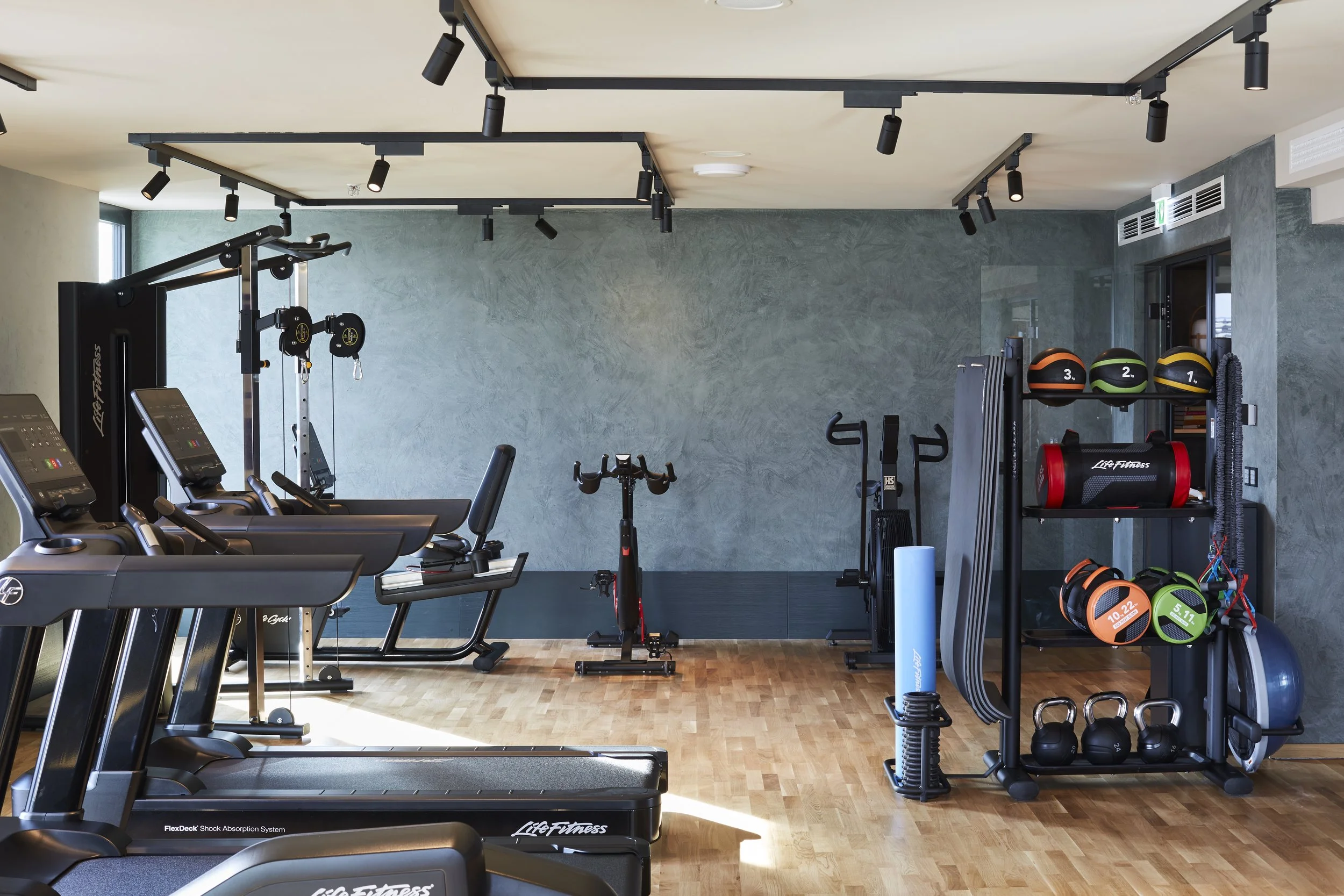 Ein Fitnessraum mit Laufbändern, Hanteln, Medizinbällen und Krafttrainingsgeräten. Die Wände sind grau, das Holzparkett ist hellbraun, und es gibt große Fenster, die Tageslicht hereinlassen.