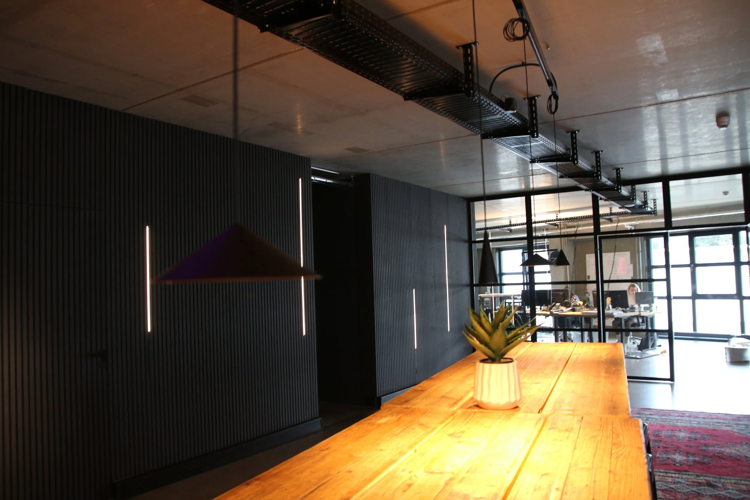 Innenansicht eines modernen Büros mit dunkler Wand, Holz-Tisch und einer stilvollen Lampe, in dem Arbeitsplätze mit Computern und Personen sichtbar sind.