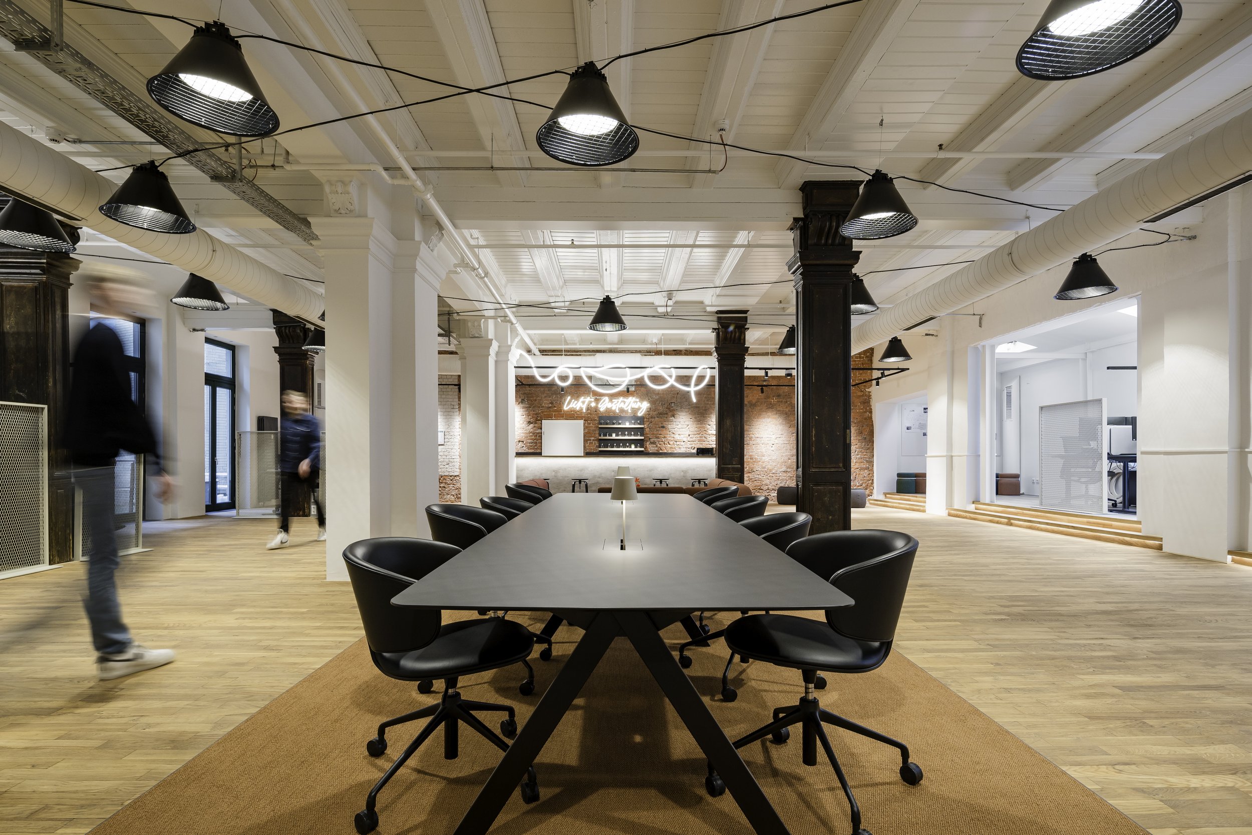 Ein moderner, heller Konferenzraum mit langem Tisch und schwarzen Stühlen, hohe Decken mit sichtbaren Holzbalken, industrielle Beleuchtung und ein Neon-Schriftzug im Hintergrund.