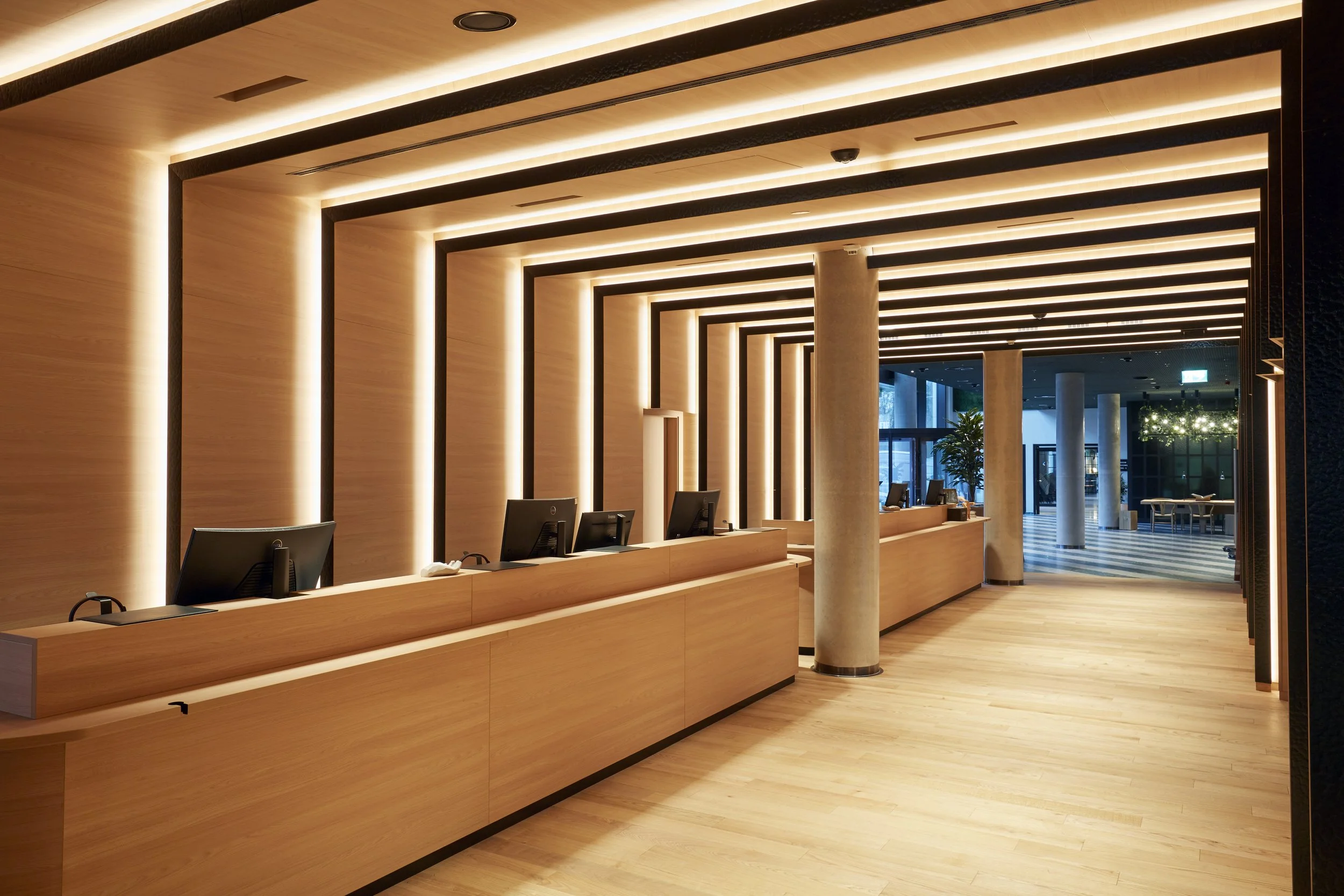 Rezeption in einem modernen Hotel mit Holztheken, Computern und moderner Beleuchtung an der Decke, im Hintergrund Fenster mit Blick nach draußen.
