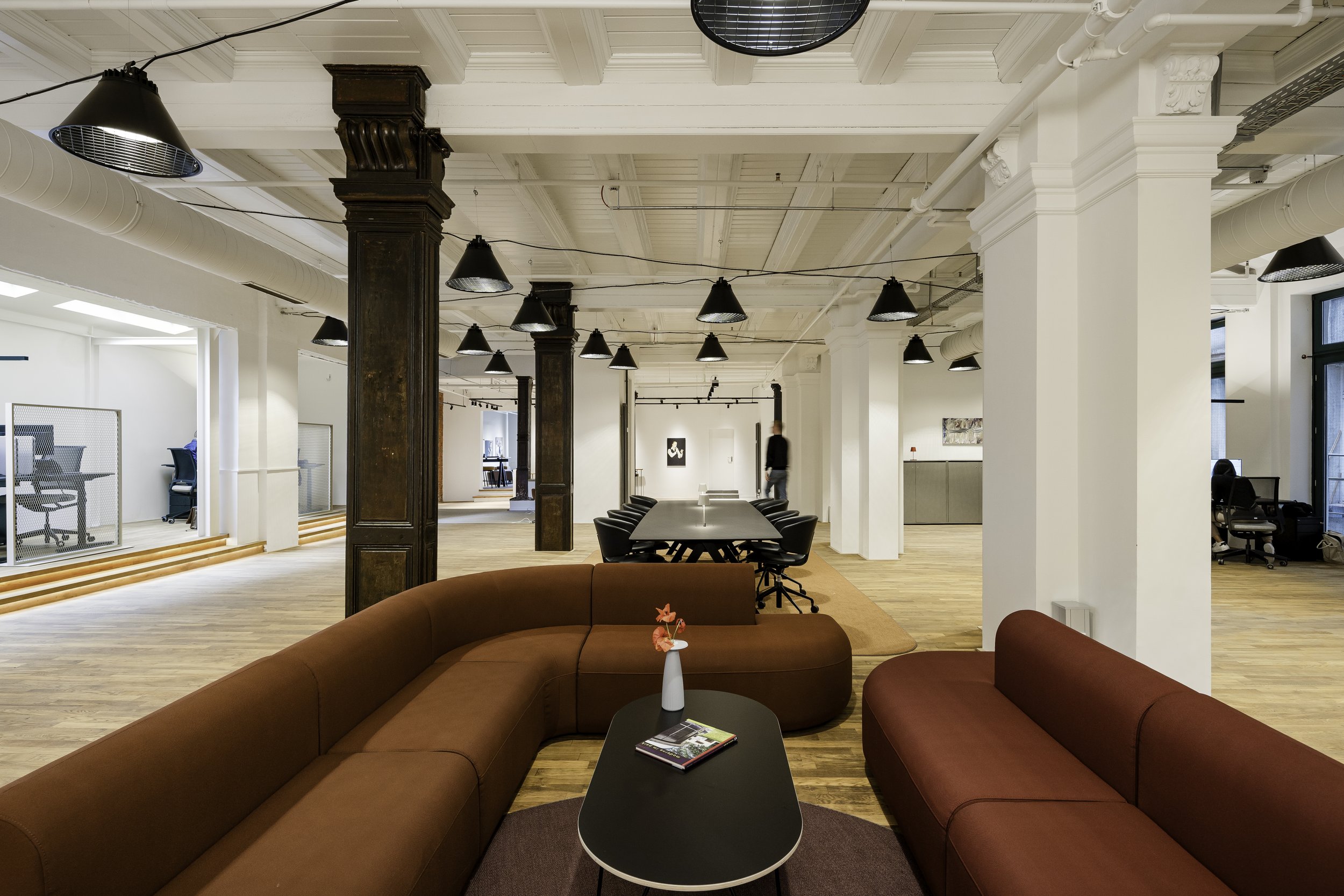 Offene, moderne Büro-Lounge mit roten Sofas, einem schwarzen Couchtisch, einer weißen Vase mit pinken Blumen, im Hintergrund helle Wände, große Fenster und weiße Decke mit schwarzen Lampen.