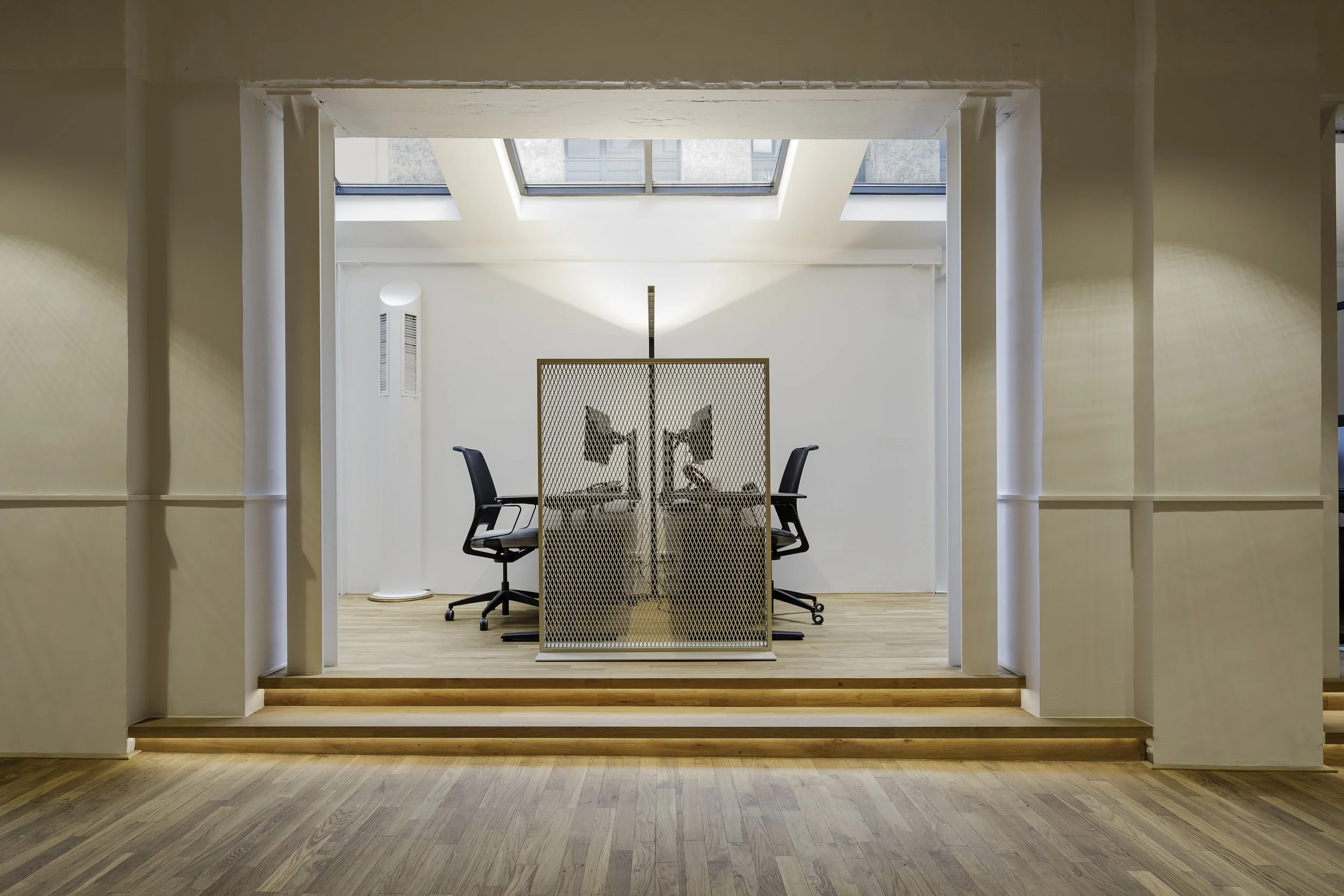 Moderner Bürobereich mit zwei Schreibtischen, Bürostühlen, einer dekorativen Trennwand, großen Dachfenstern und natürlichem Licht in einer hellen, minimalistisch gestalteten Umgebung.