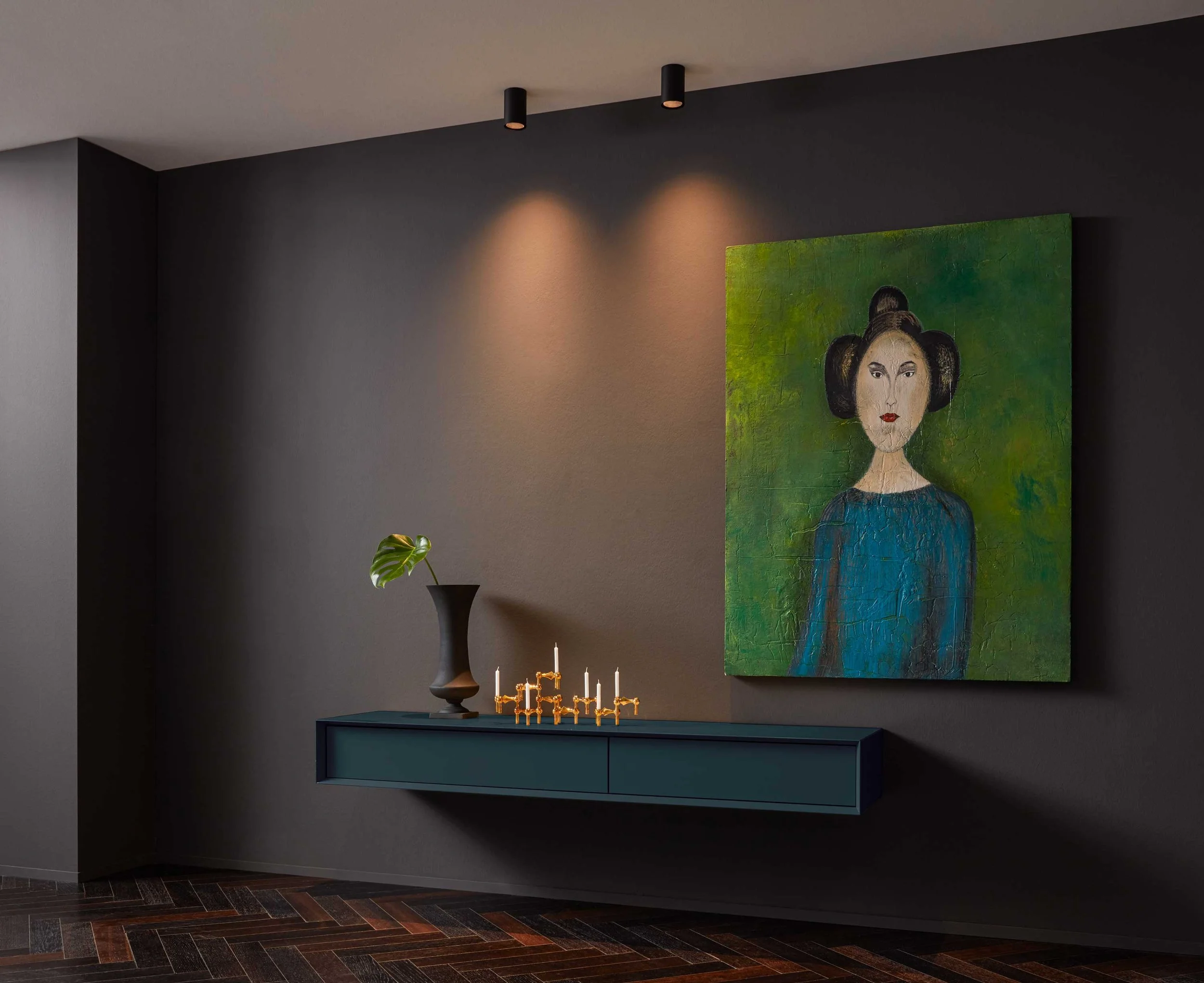 Eine dunkle Wand mit einer Darstellung eines weiblichen Porträts in einem grünen Hintergrund. Darunter ein schmaler blauer Wandtisch mit einem schwarzen Vaset, in dem eine grüne Monstera-Pflanze steht. An der Wand hängen zwei schwarze Deckenstrahler,