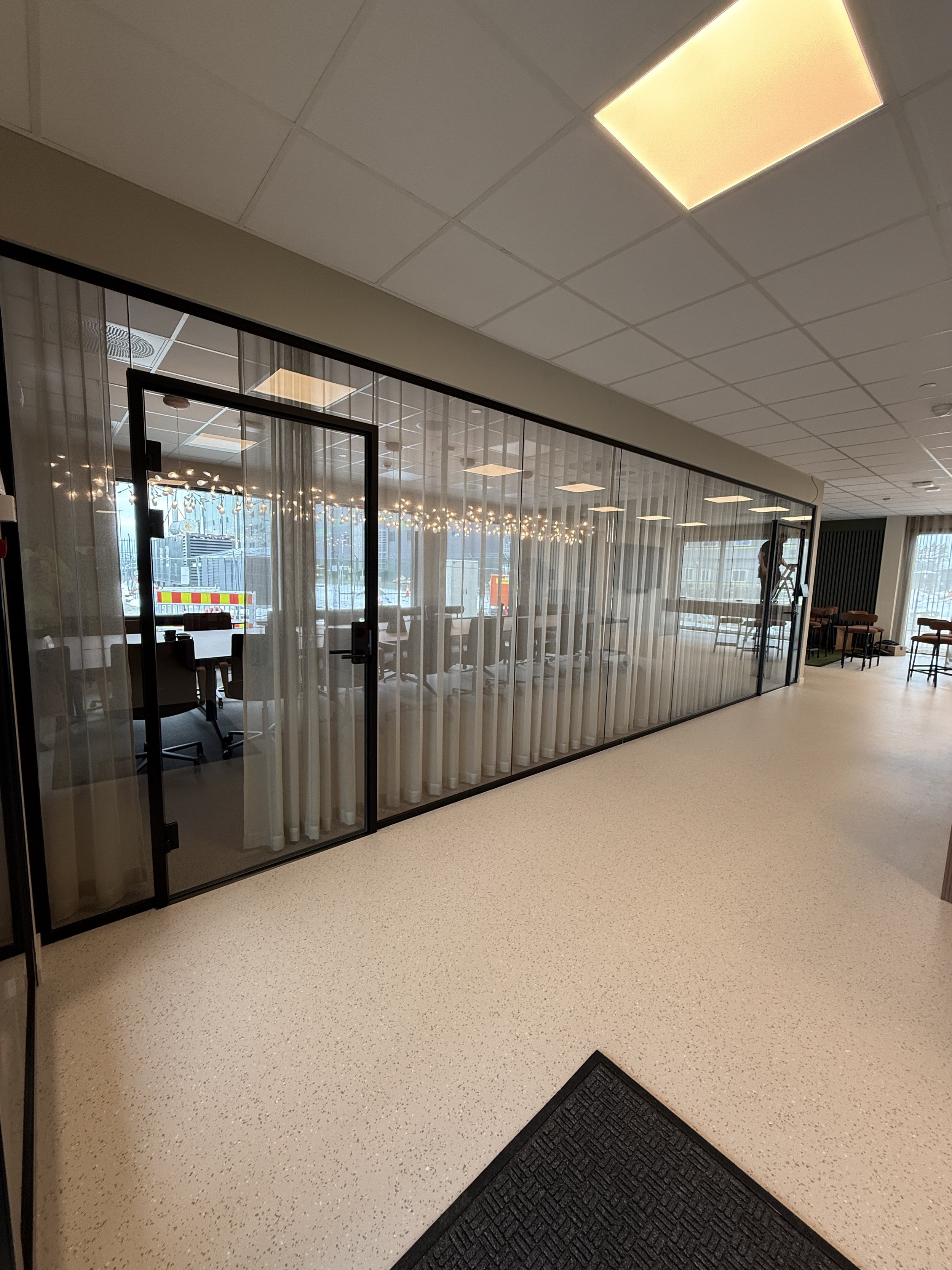 Innendørs utsikt av kontorlokaler med glassvegg, layert lys på taket, og tilstøtende områder med stoler og bord.