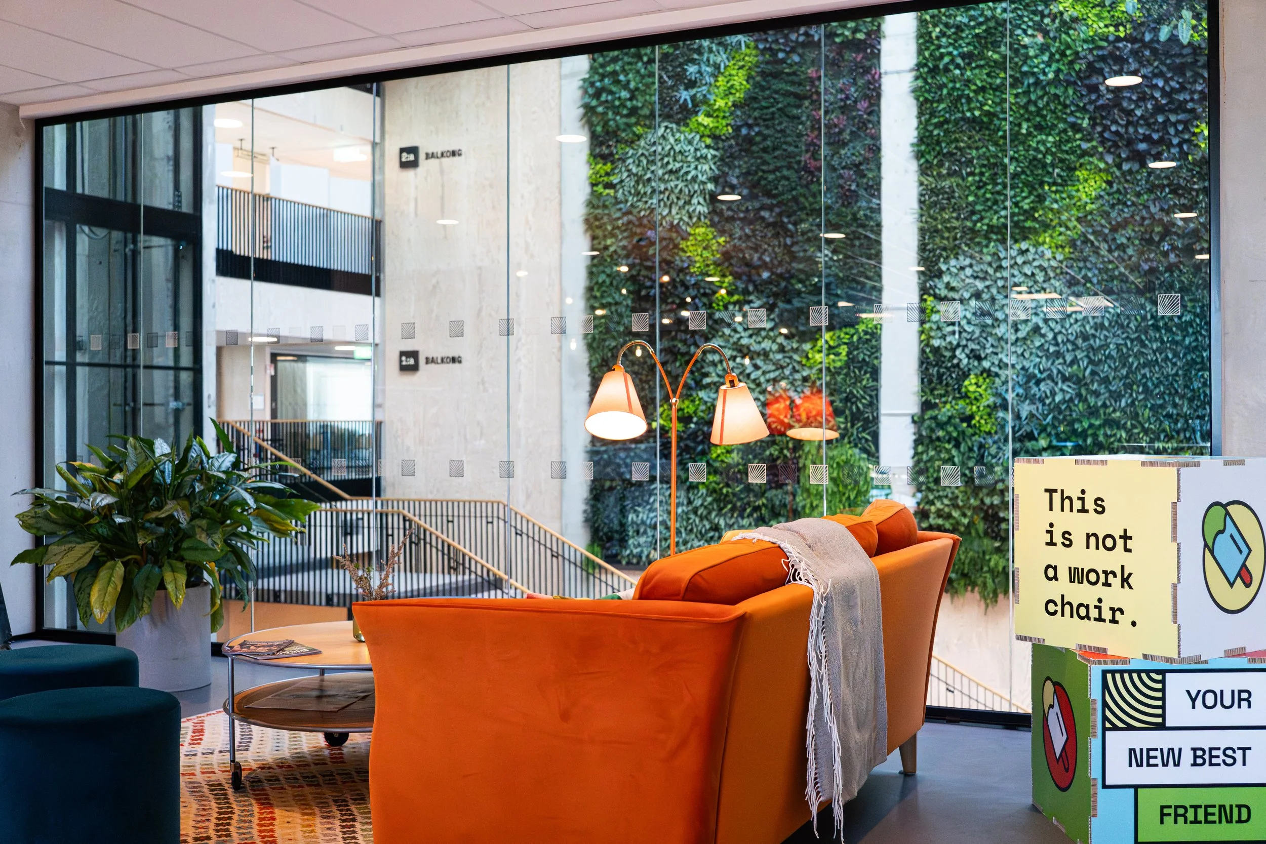 Innendørsområde med oransje sofa, grønn plante, bord med magasiner, og en stor glassvegg med utsikt til en grønn vegg utenfor.