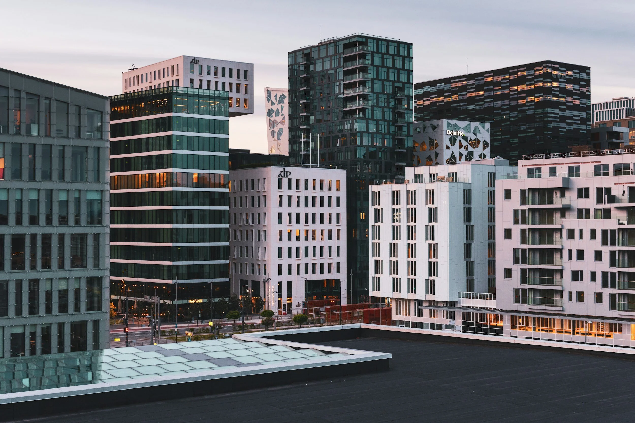 Utsikt over moderne kontorbygninger i et bymiljø med mørke og lyse fasader og ettermiddag- eller kveldsskyer.