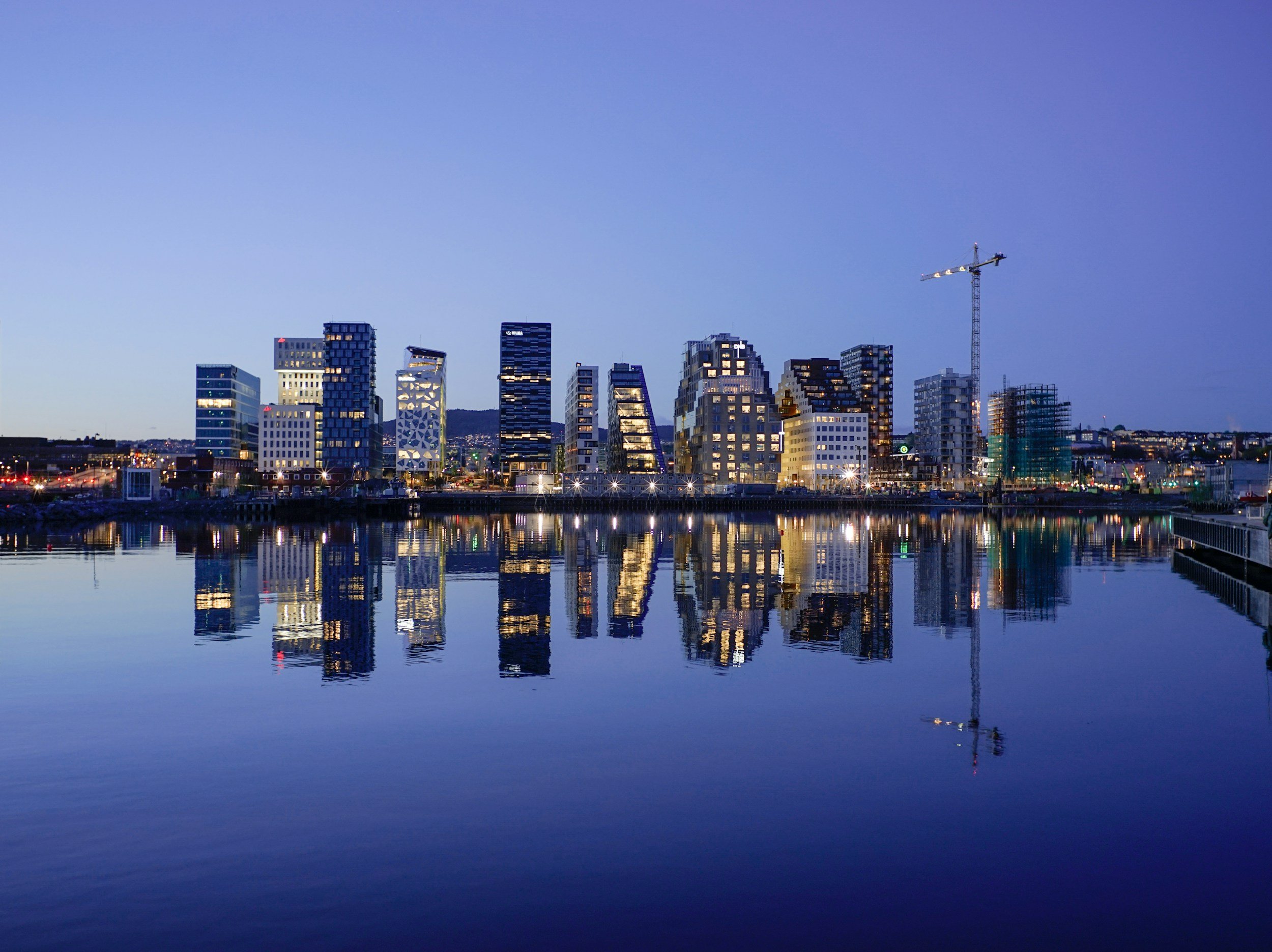 Utsikt over Oslo, by med moderne høyhus speilet i vannet ved elvebredden om kvelden. Bygninger er opplyst, og det er en kran på høyden, som indikerer byggeaktivitet.