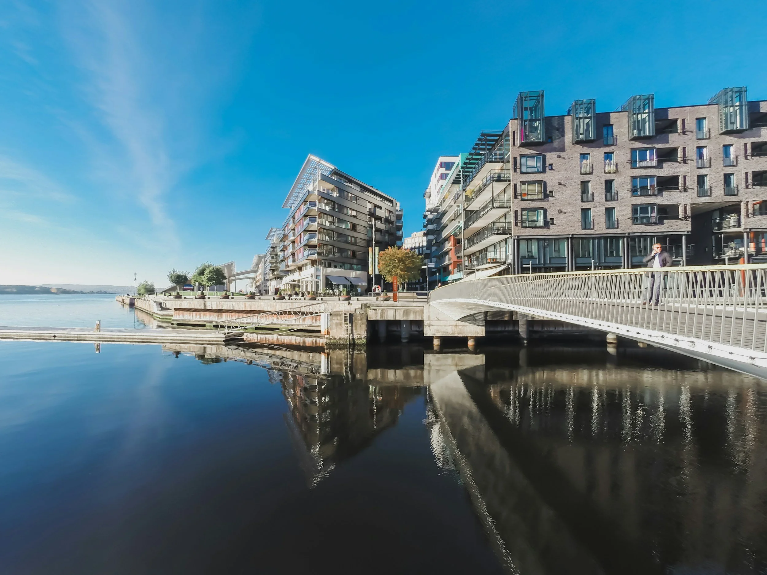 Moderne bygninger ved Aker Brygge i Oslo, ved vannkanten med refleksjon i vannet, klar blå himmel og en person som går og snakker i telefon.